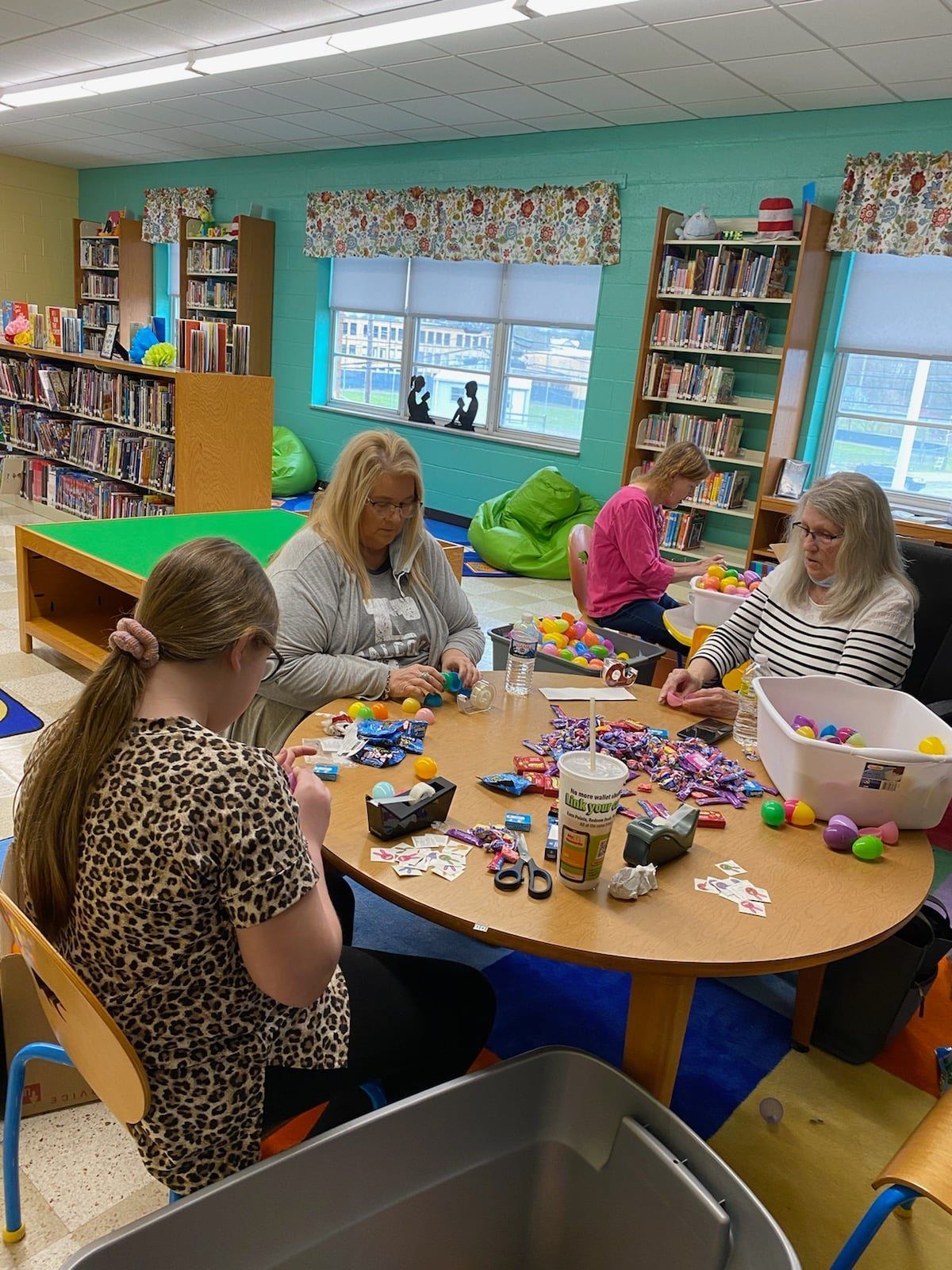 People seated around a table sorting small items, likely in a library. Bookshelves are visible in the background.