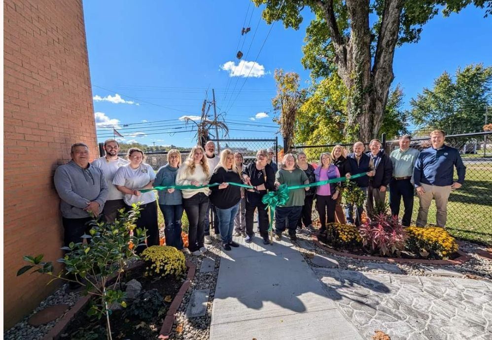 Group of people at a ribbon-cutting ceremony outdoors near a brick building and a tree.
