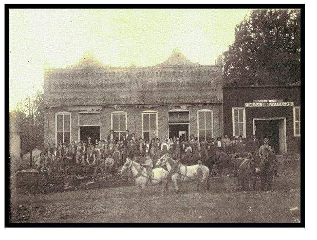 Old photo: Group of people outside a two-story building. Several men on horseback in front.