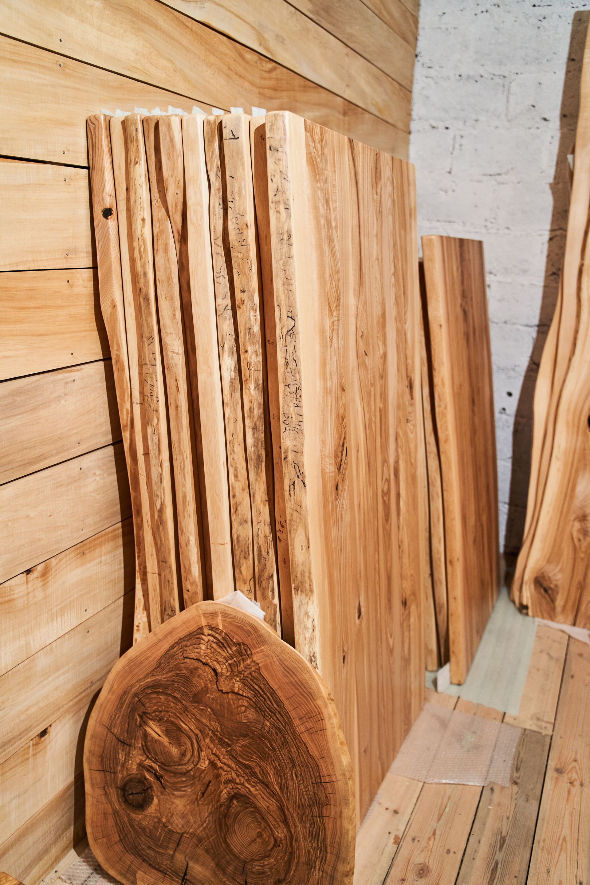 Stack of raw wooden slabs in a workshop, with live edges and visible wood grain, against a wooden wall.