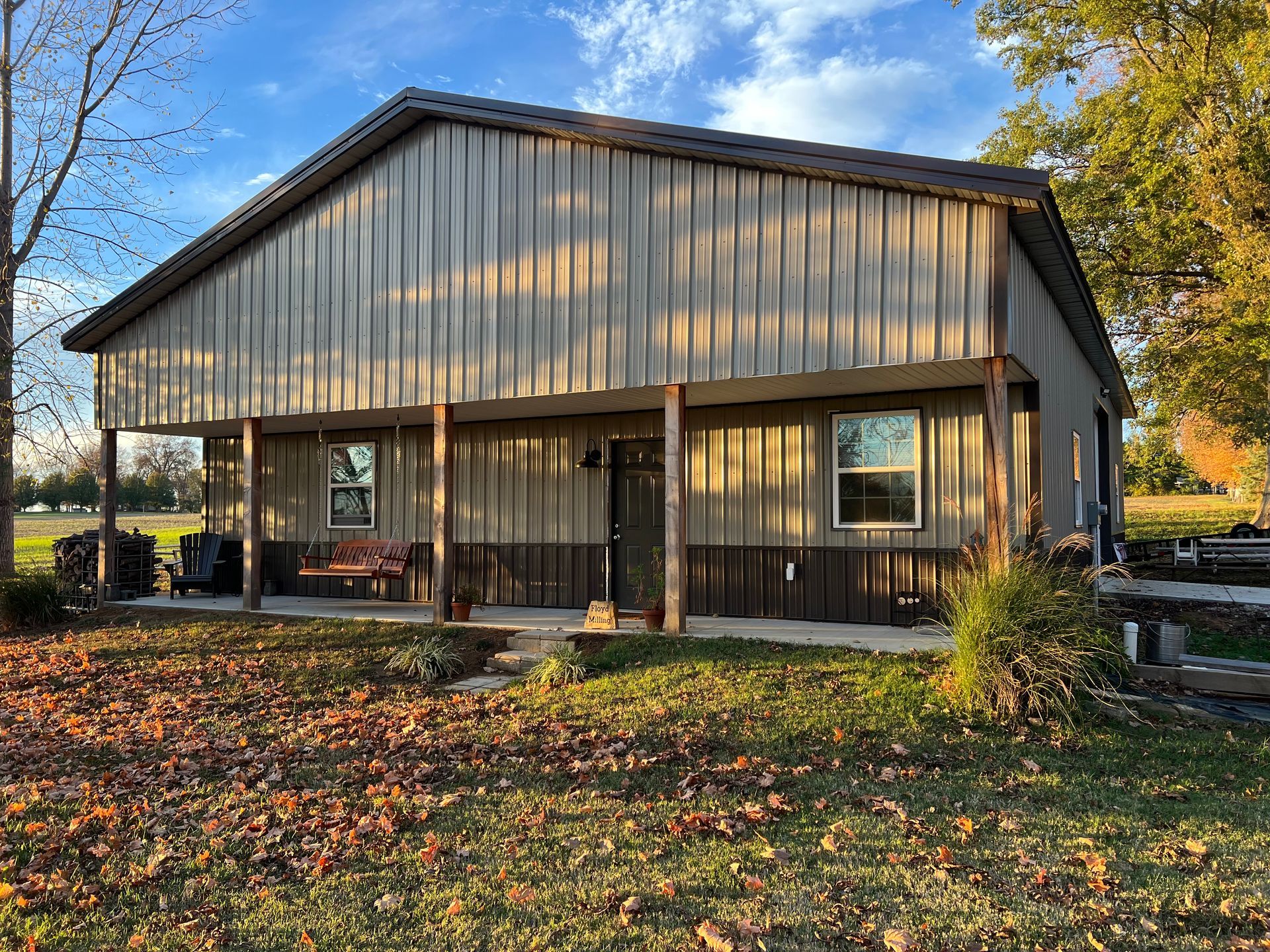Two-tone metal-sided building with porch, windows, and brown columns on a grassy field with fallen leaves.