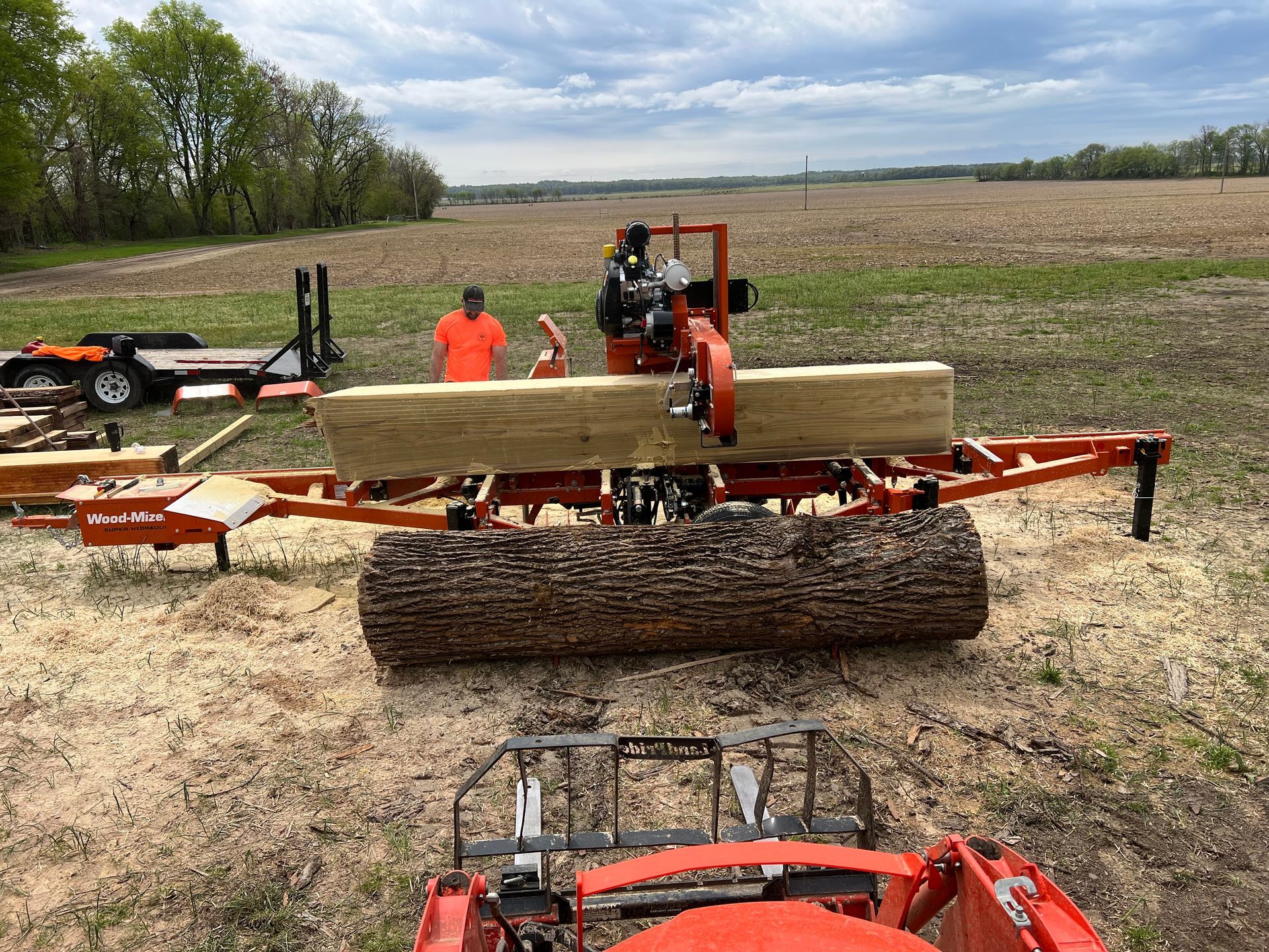 Person operating a sawmill, cutting a log in a field. Tractor in foreground, trailer in background.