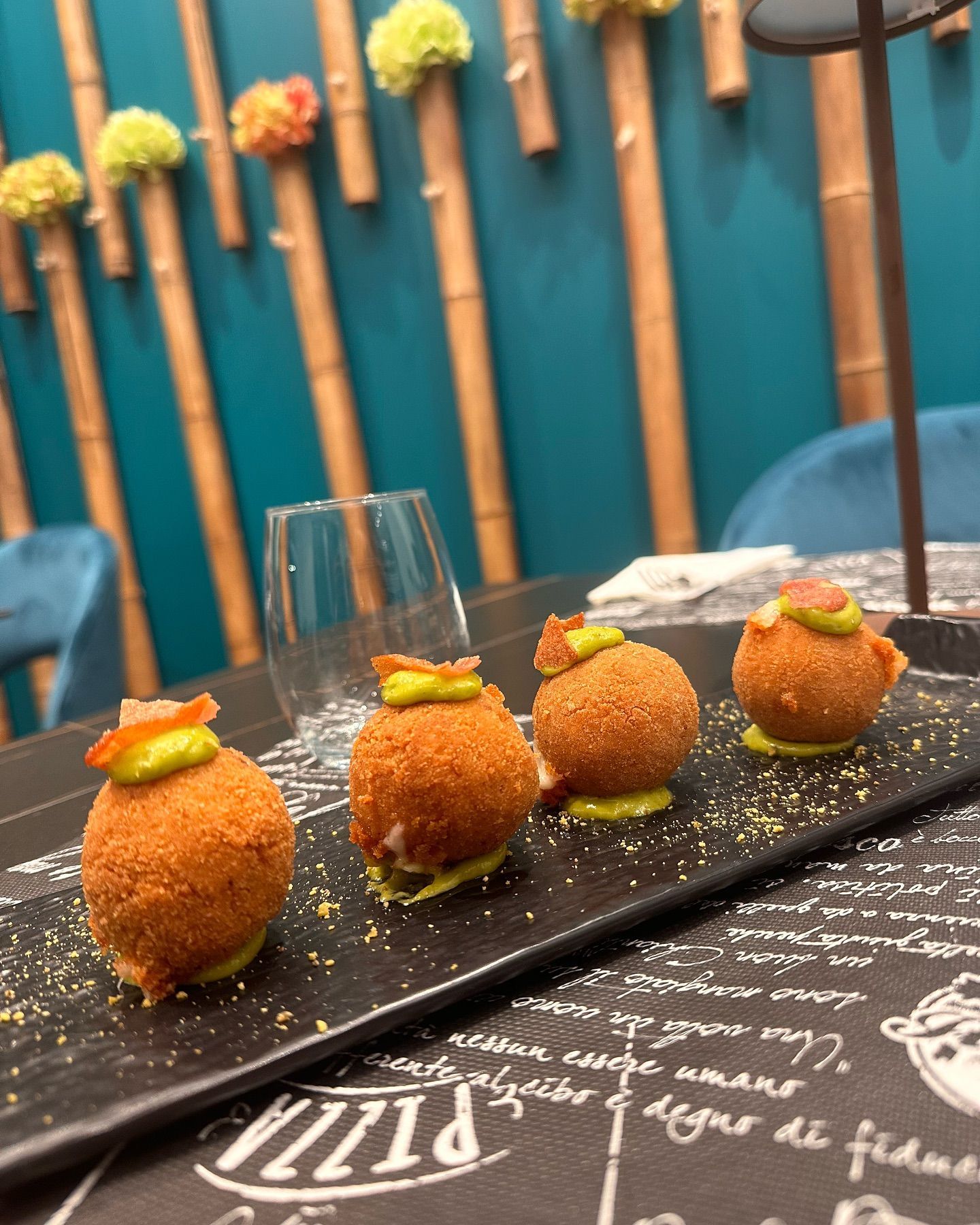 Quattro antipasti rotondi e dorati con salsa verde su un piatto nero. Sfondo turchese con bambù.