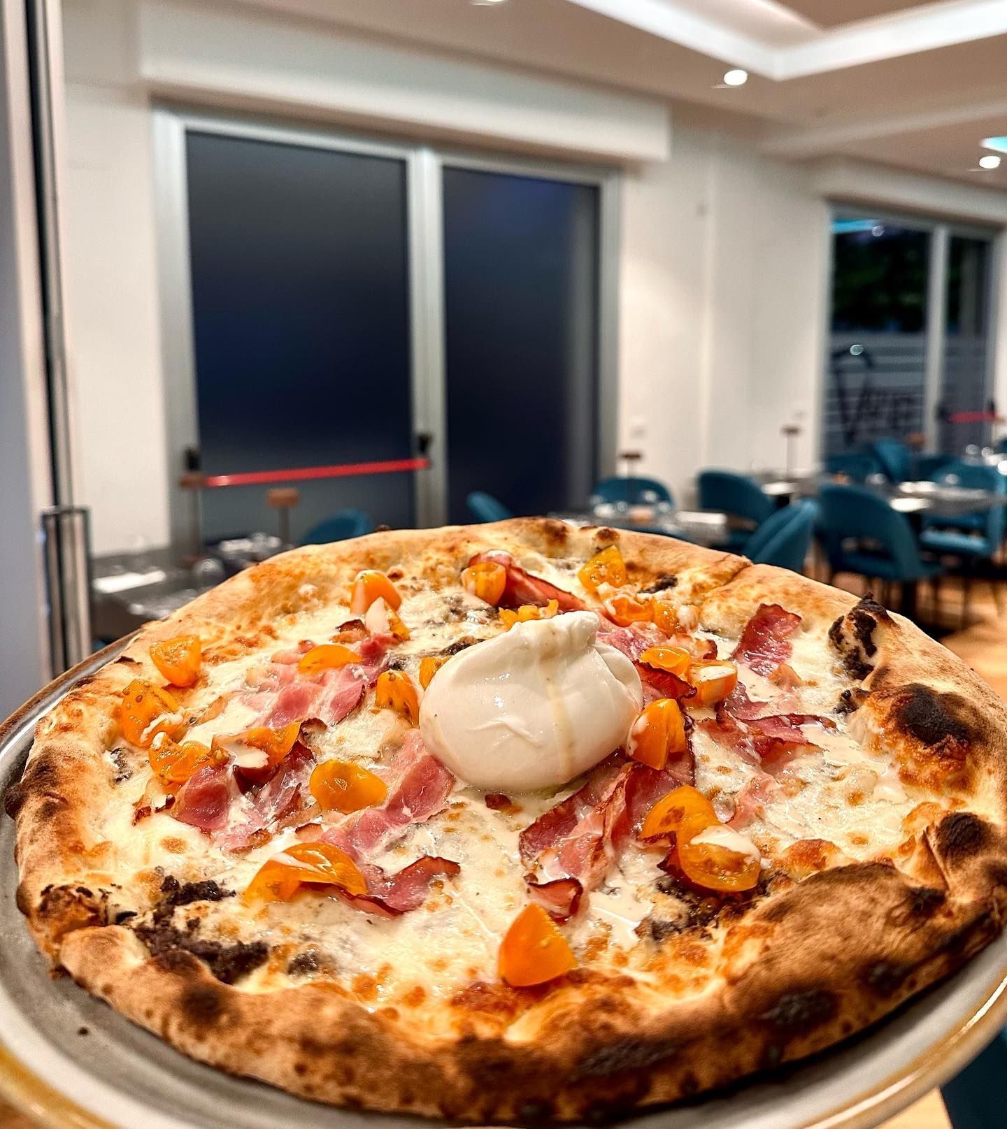 Pizza su un vassoio; condita con formaggio, pancetta e pomodori. Sullo sfondo, l'interno del ristorante.