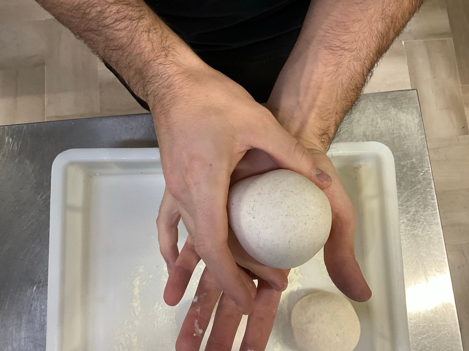 Mani che tengono e danno forma a una palla di impasto, un'altra palla di impasto in un vassoio, probabilmente per fare la pizza.