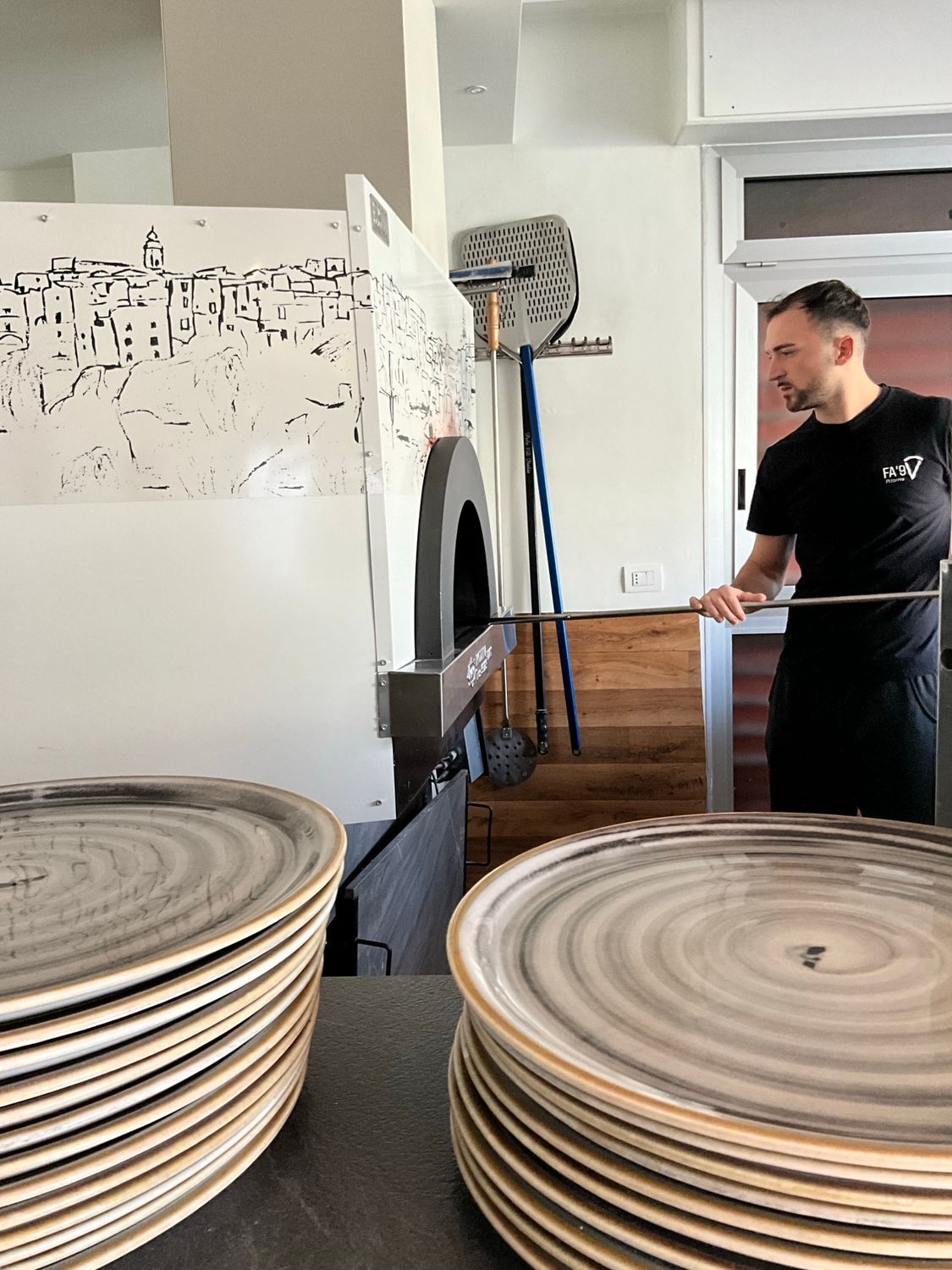 Uno chef che si occupa di un forno per pizza. Pile di piatti in primo piano. Interno ben illuminato.