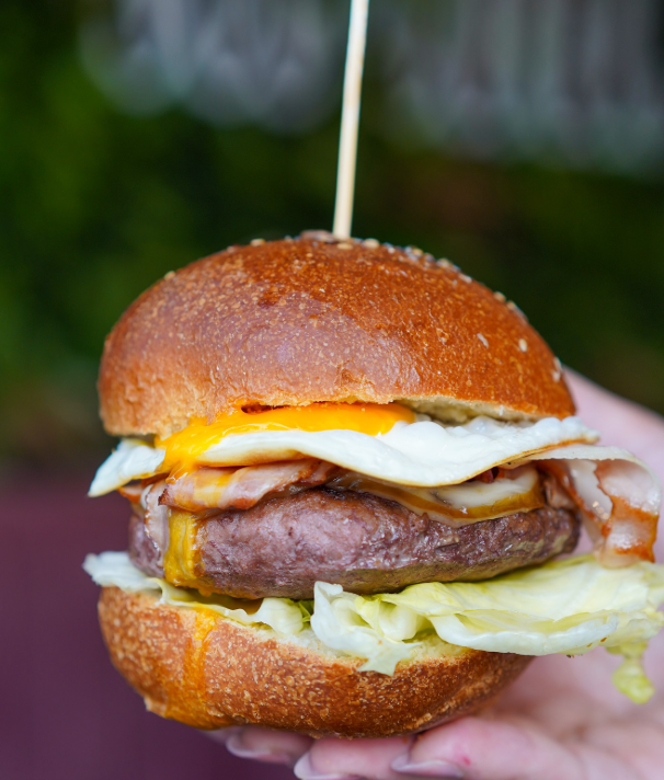 Hamburger con uovo, pancetta e lattuga su un panino, tenuto in mano e fissato con uno stuzzicadenti.