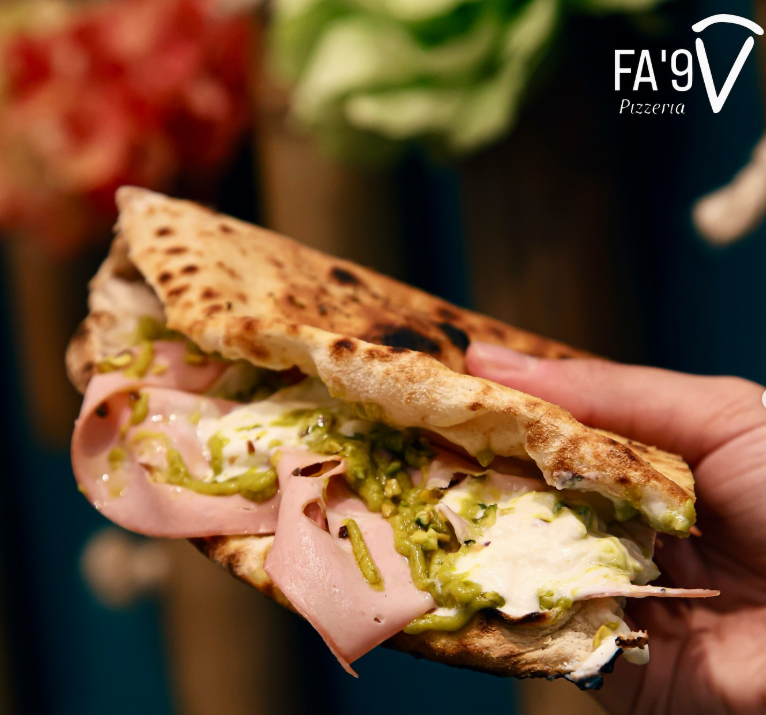 Hand holding a pizza sandwich with mortadella, pesto, and mozzarella. FA'9 Pizza logo in corner.