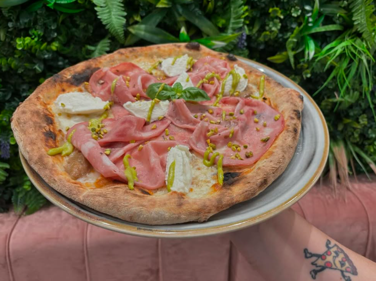 Pizza con mortadella, mozzarella e pistacchi su un piatto, tenuta da una persona con un tatuaggio.