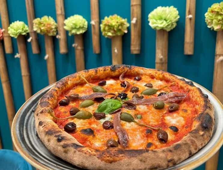 Pizza con olive, capperi, acciughe e basilico su un piatto. Appoggiata a una parete turchese con decorazioni in bambù.