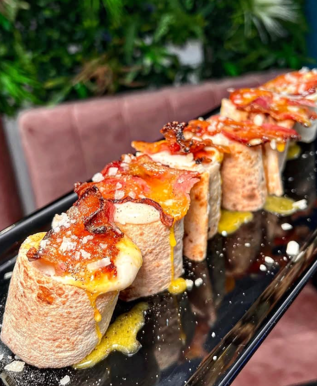 Panini in stile sushi conditi con pancetta e formaggio, conditi con salsa e serviti su un piatto nero.