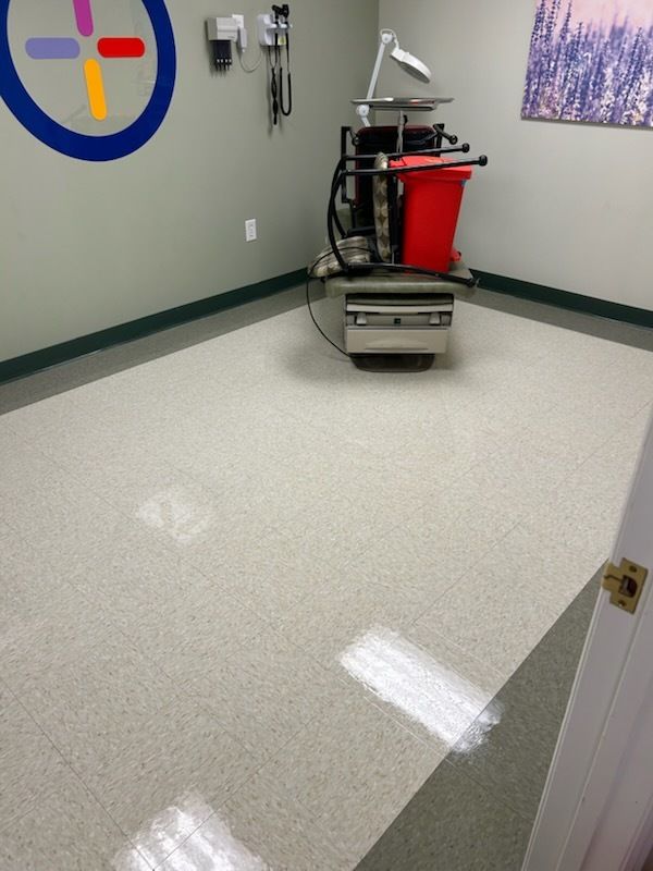 A view into a clinical examination room with light-colored flooring, a medical cart, a red waste bin, and wall equipment.