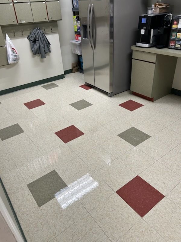 An office break room with white walls, cabinets, a stainless steel refrigerator, coffee maker, and a tiled floor.