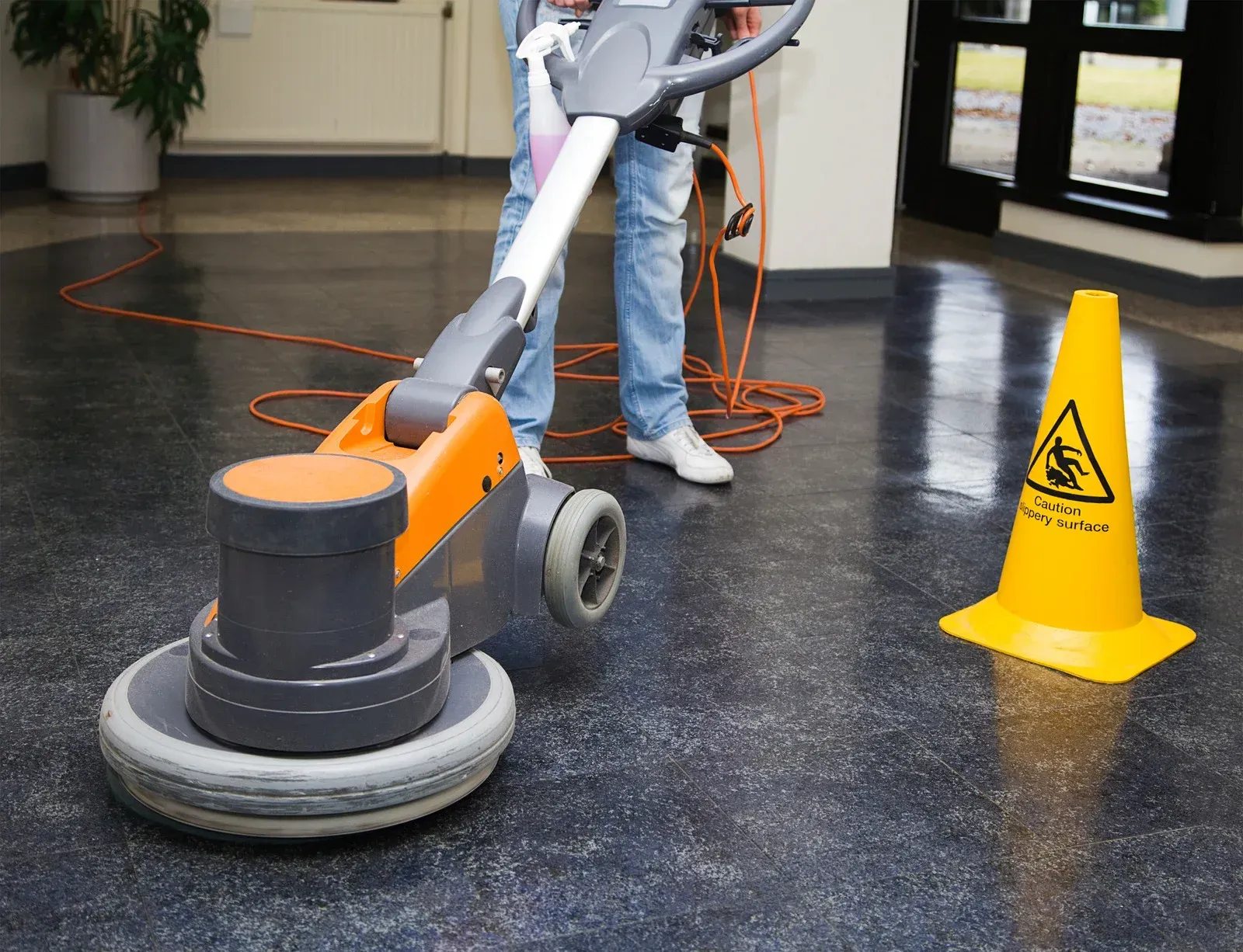 A person is using a machine to clean a floor next to a yellow caution cone