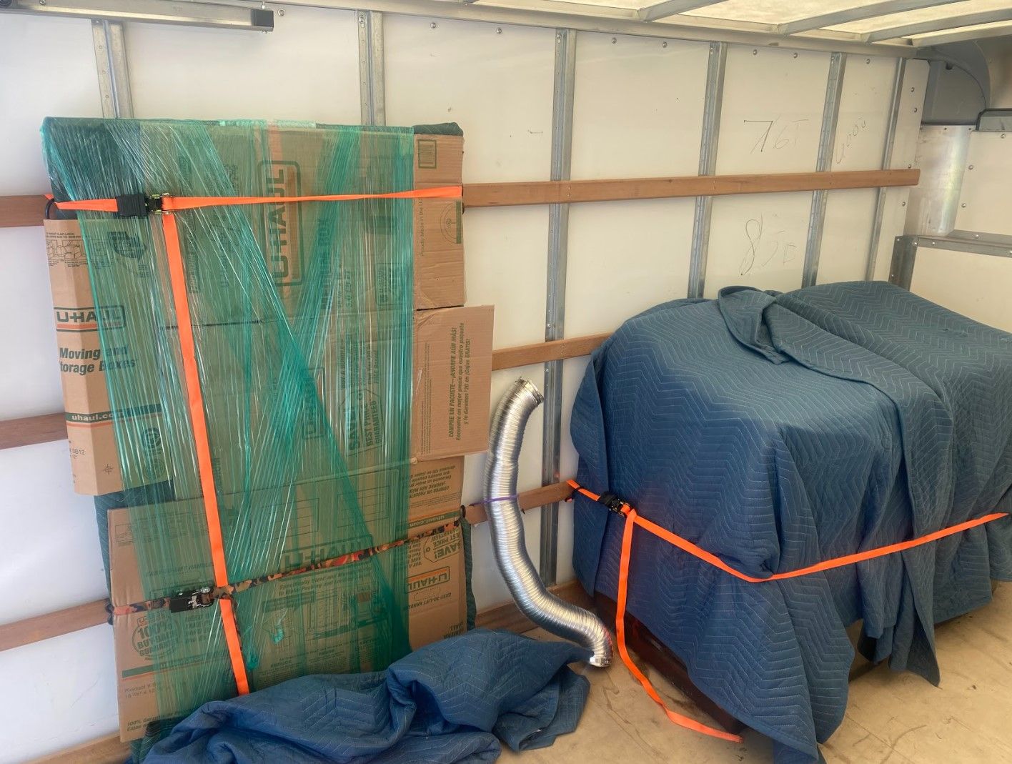 Inside a moving truck, boxes and furniture wrapped in blue pads secured with orange straps.
