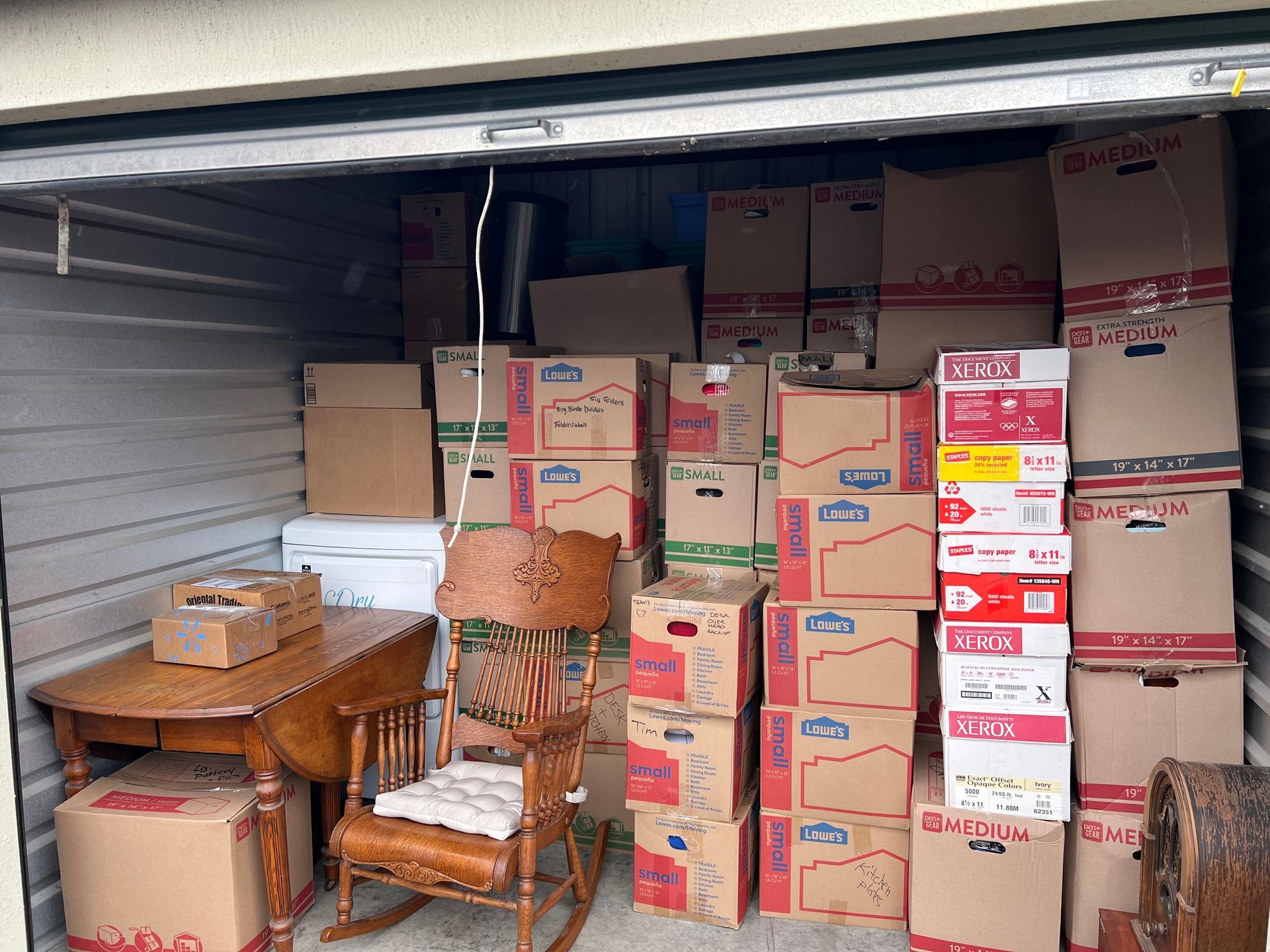 Storage unit packed with cardboard boxes, a wooden table, rocking chair, and other furniture.