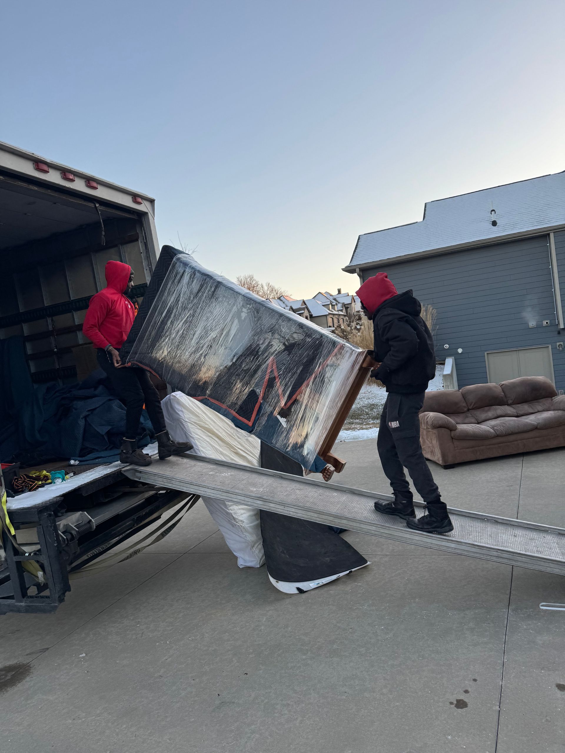 Two people loading a large, wrapped furniture item onto a truck ramp outside. Cold, overcast setting.