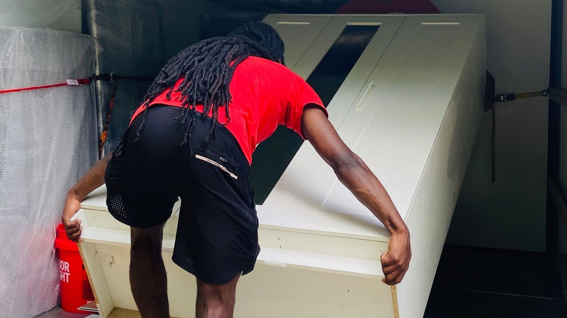 Person in red shirt and black shorts lifting a white cabinet inside a truck.