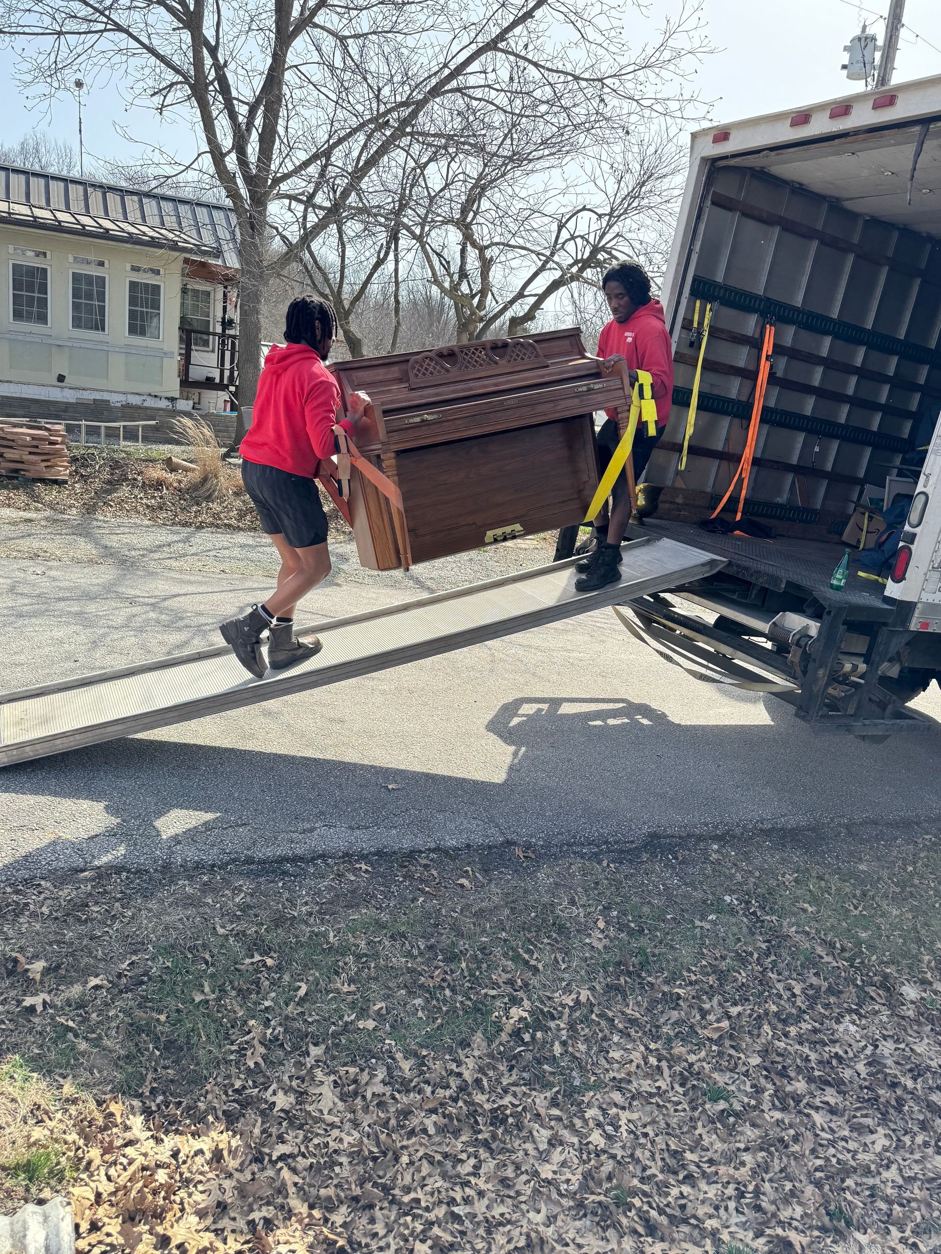 Two movers in red shirts load a small piano onto a truck on a sunny day.
