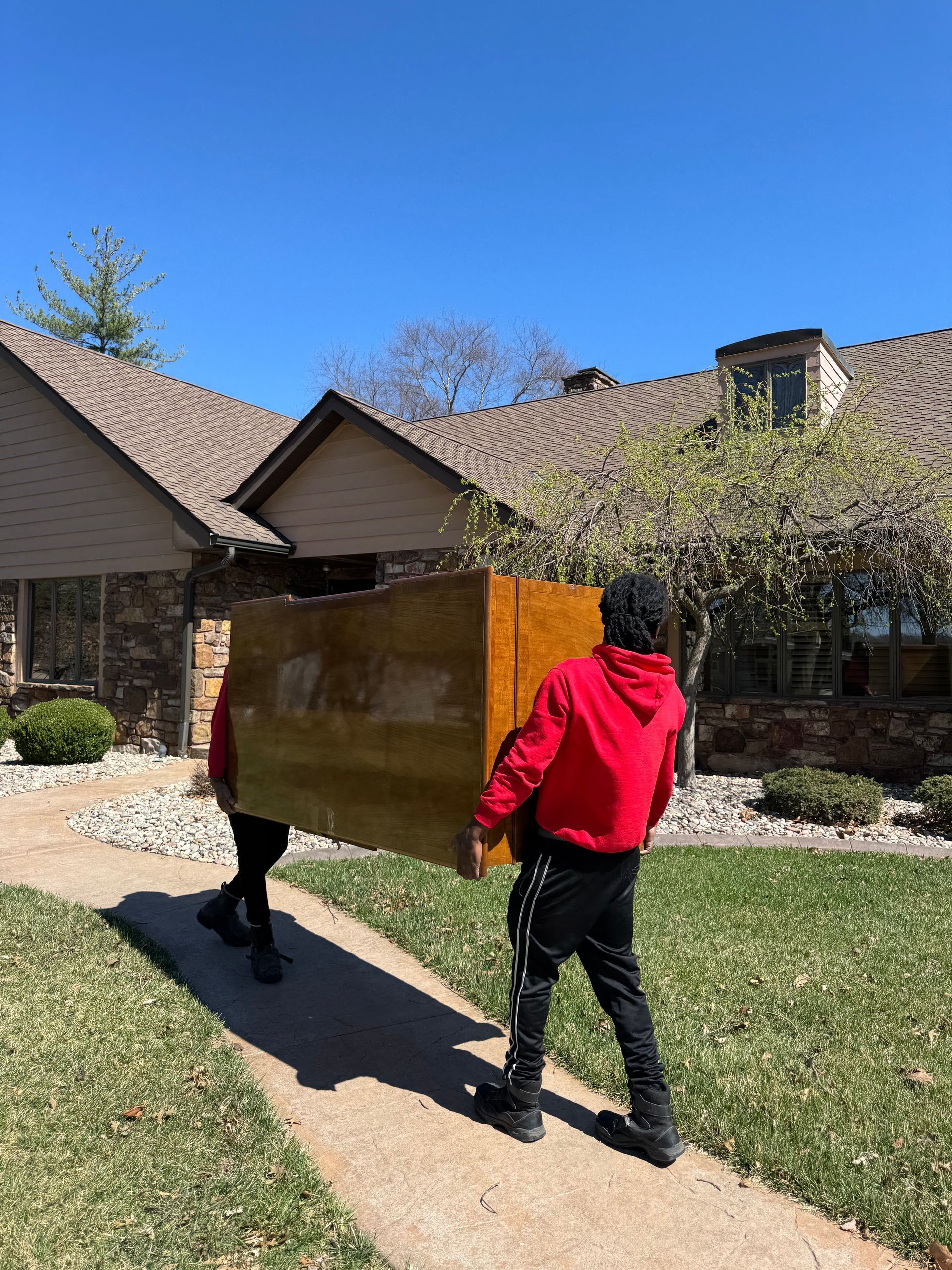 Two people carrying a large, light-colored wooden object down a sidewalk toward a house on a sunny day.