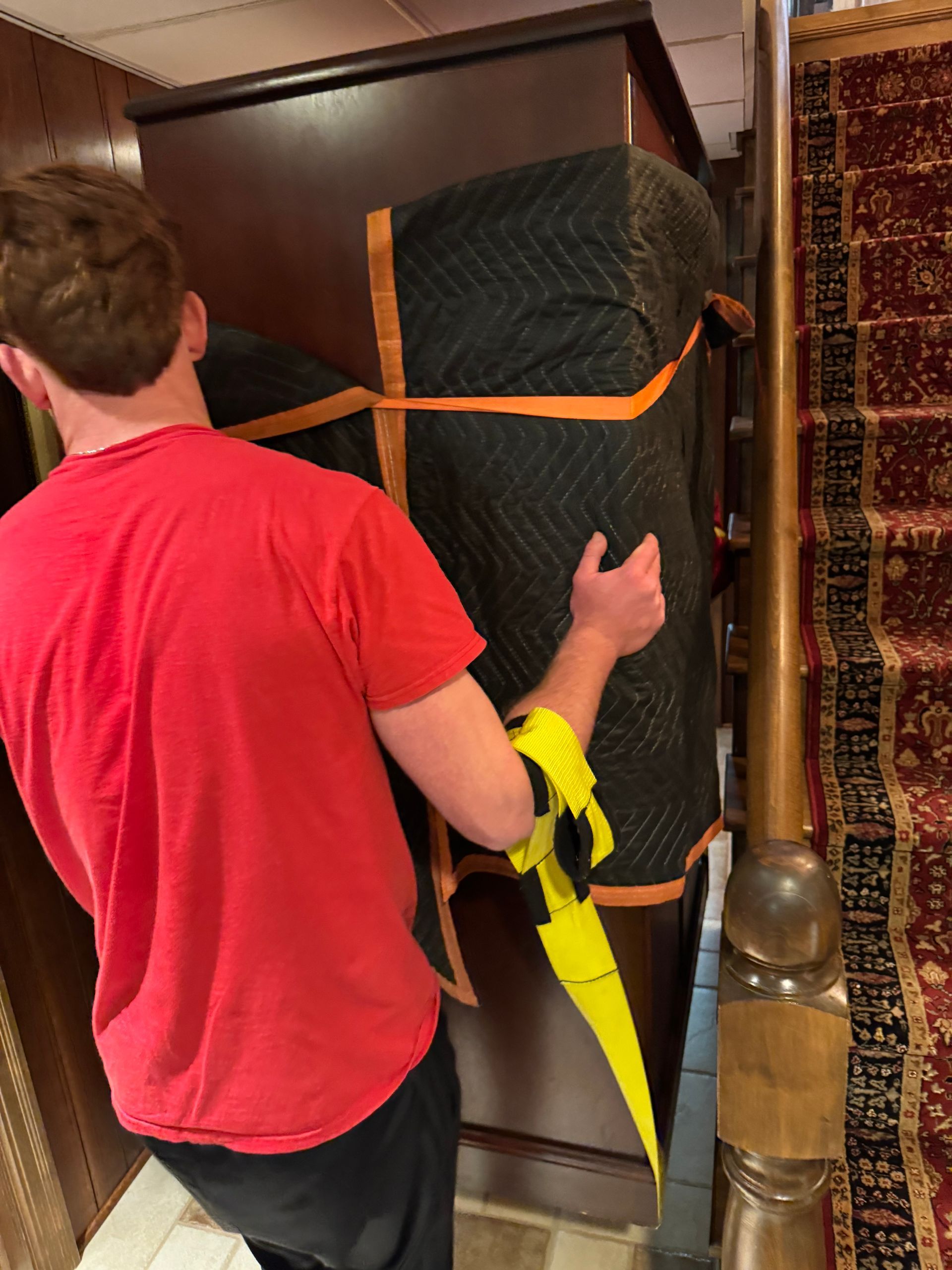 Man in red shirt carrying a wrapped, tall piece of furniture down a staircase, using straps for support.