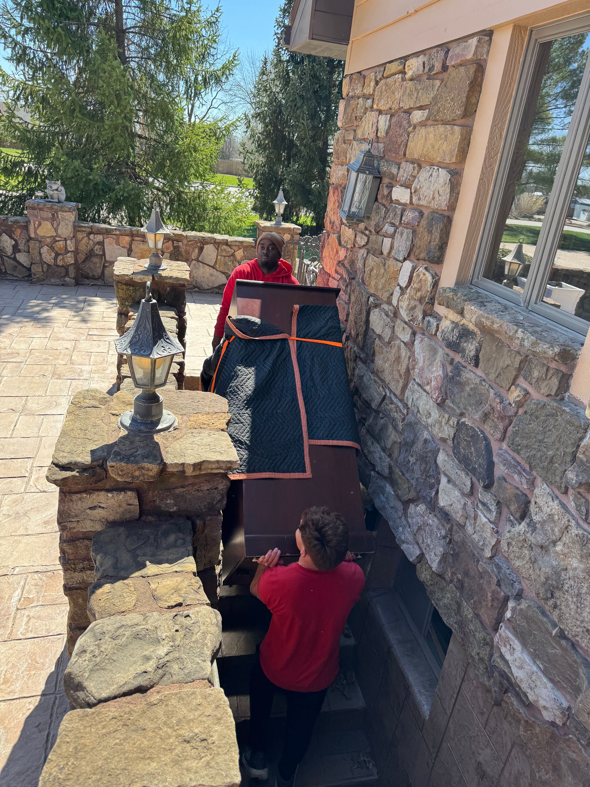 Two people in red shirts moving a large wooden object near a stone building.