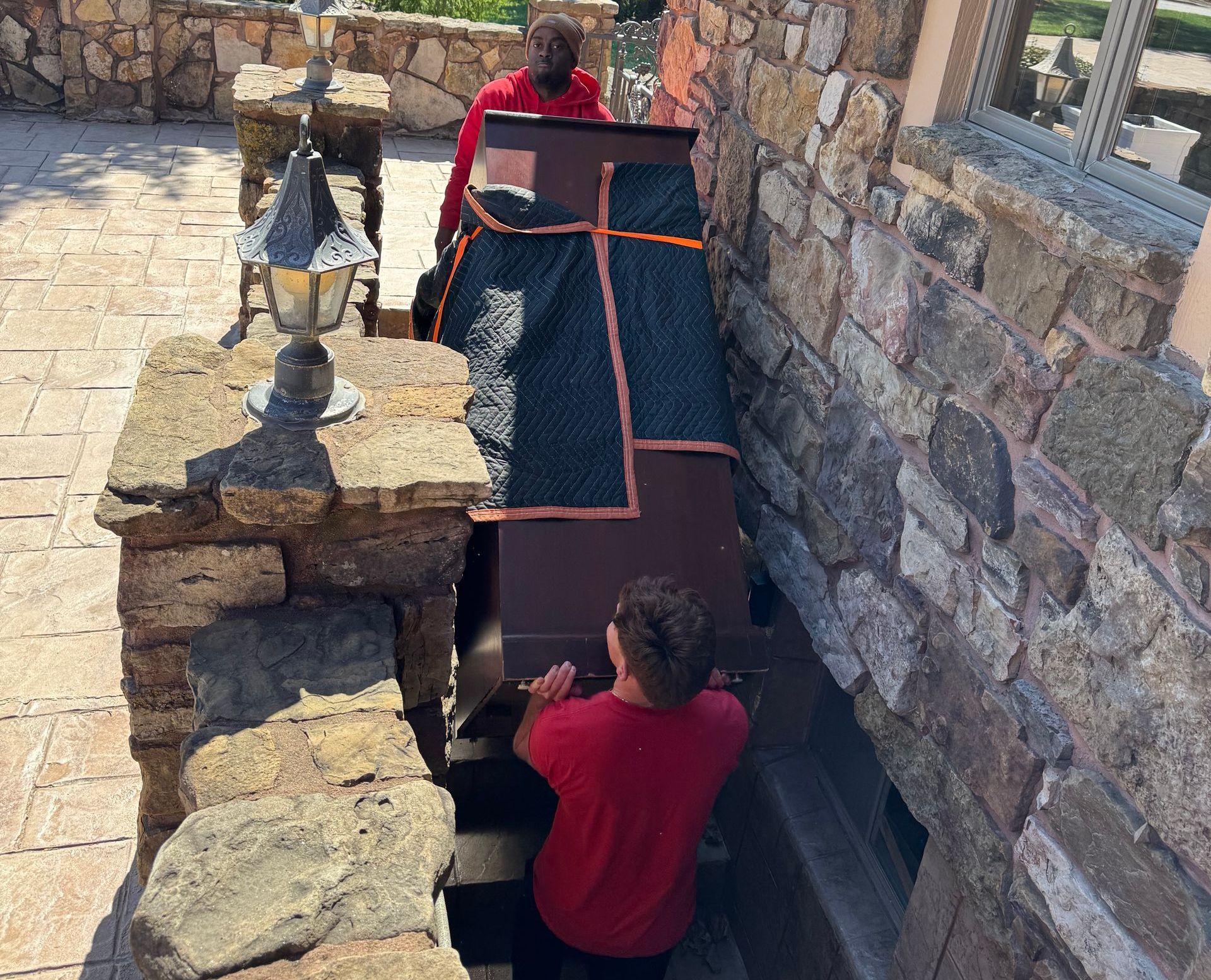 Two people moving a wrapped brown object up stone stairs. Red shirts, outdoor setting.