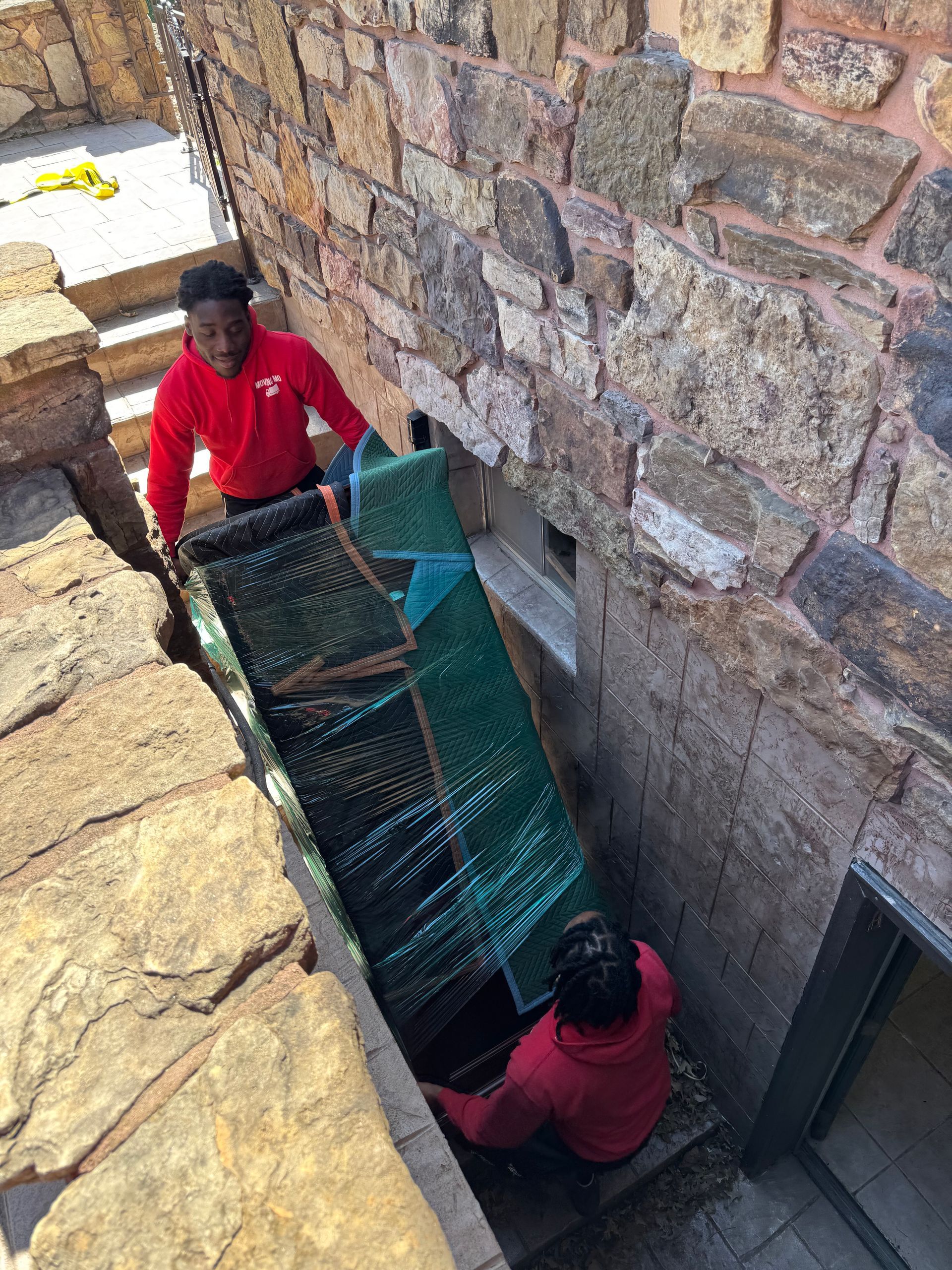 Two men in red shirts carry a long, green-wrapped object through a narrow opening in a stone wall, down steps.