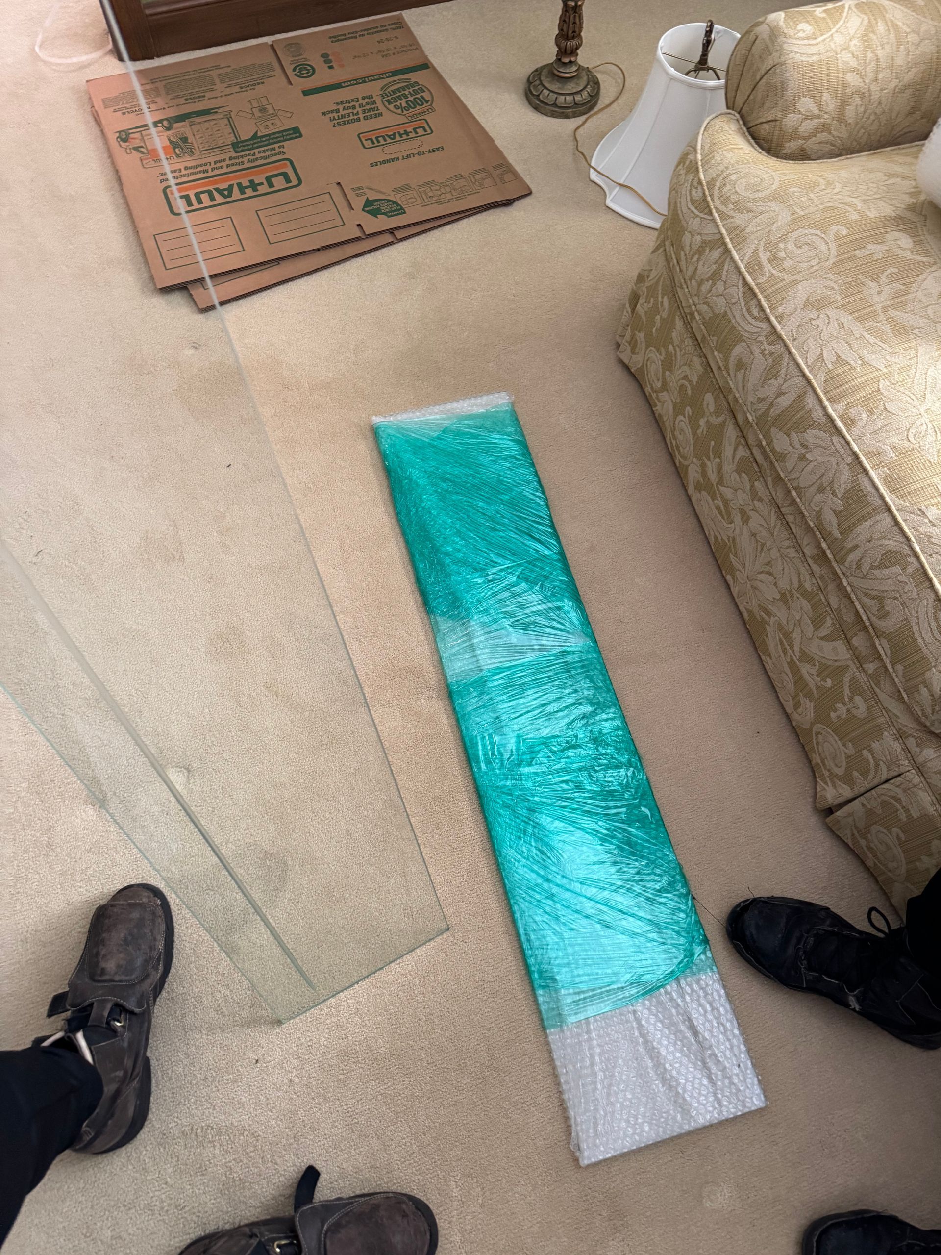 A room with carpet, glass, bubble wrap, moving boxes, and furniture. Two pairs of shoes are visible.