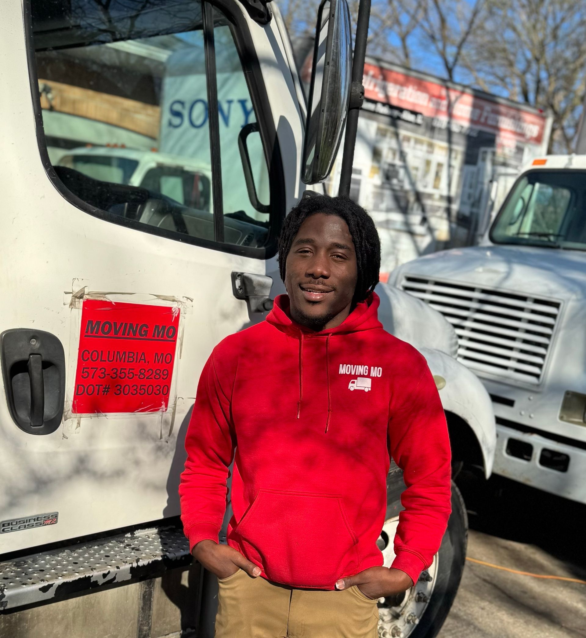 Man in red hoodie, standing in front of a truck. Outdoors, sunny. Brown pants, hands in pockets.