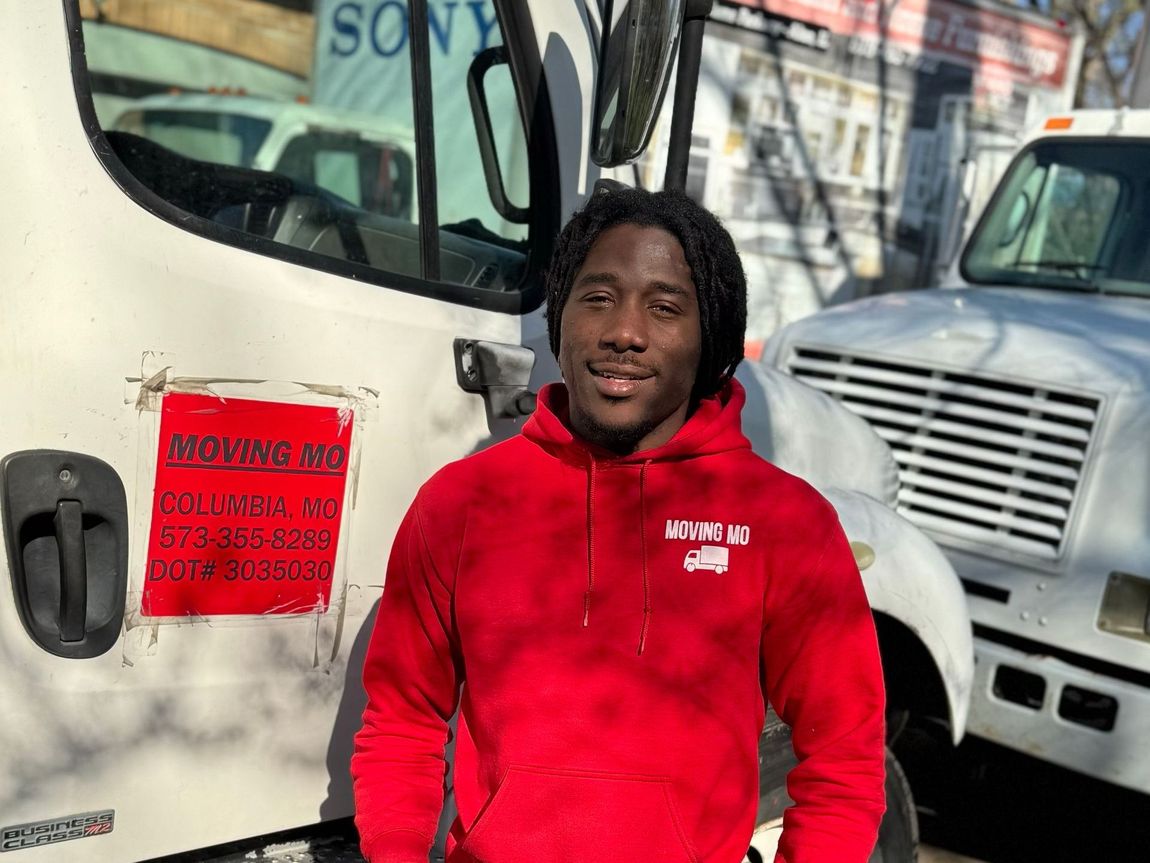 Man in red hoodie stands by white trucks; sunlight, outdoor setting.
