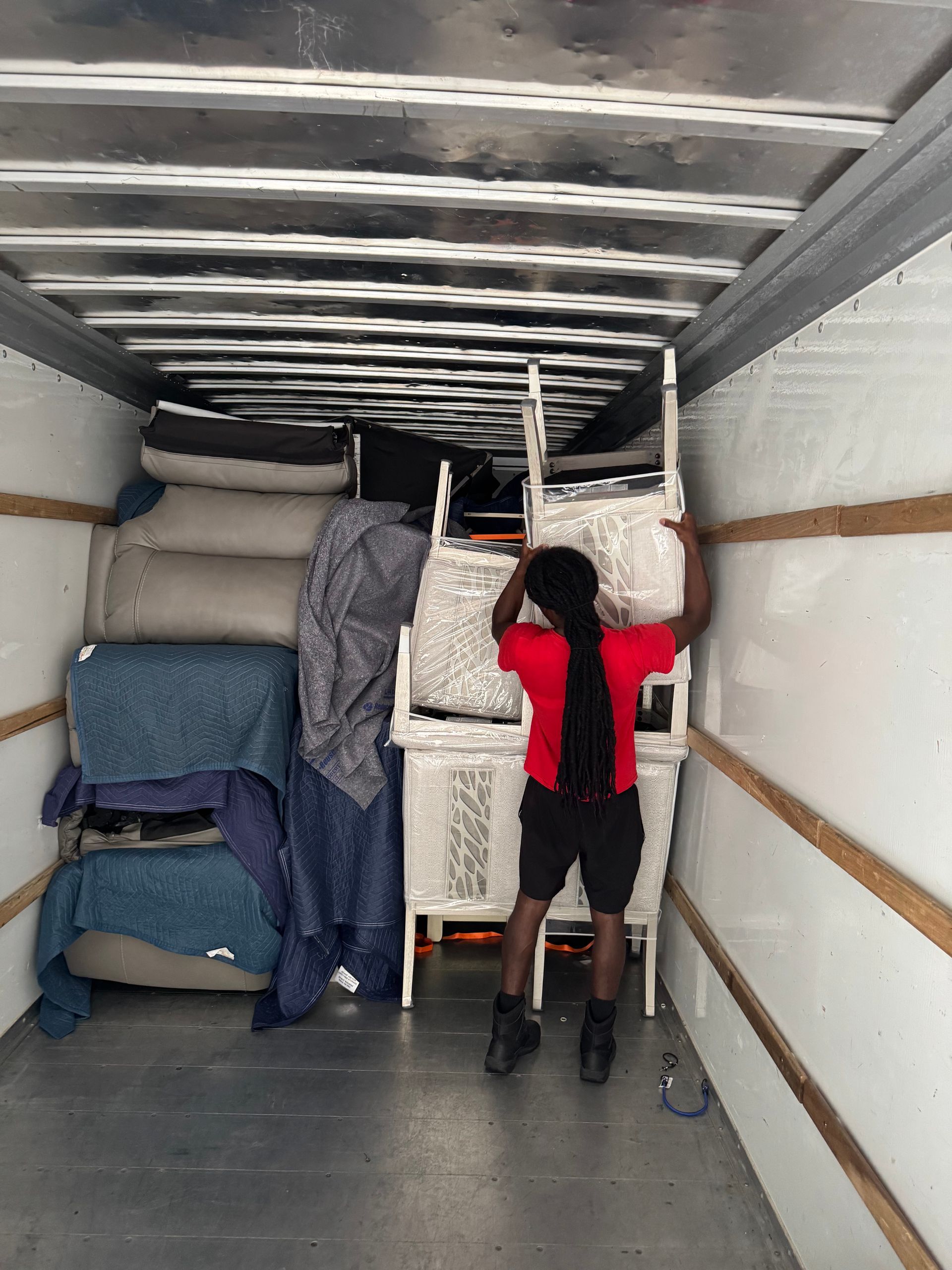 Person in a red shirt inside a moving truck, holding a white dresser. Packed furniture fills the truck.
