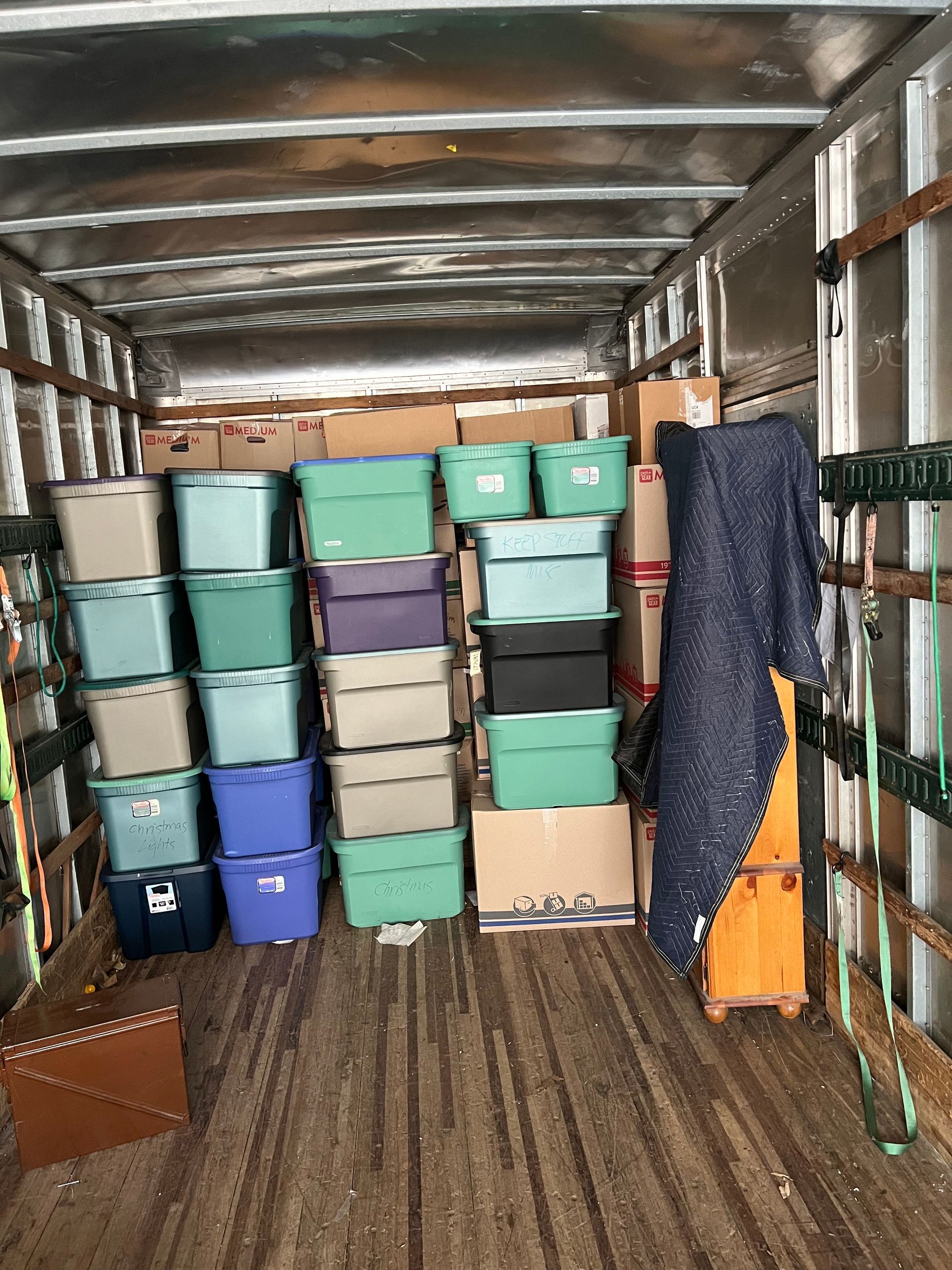 Inside a moving truck: boxes and bins stacked, with luggage and a blanket on the right.