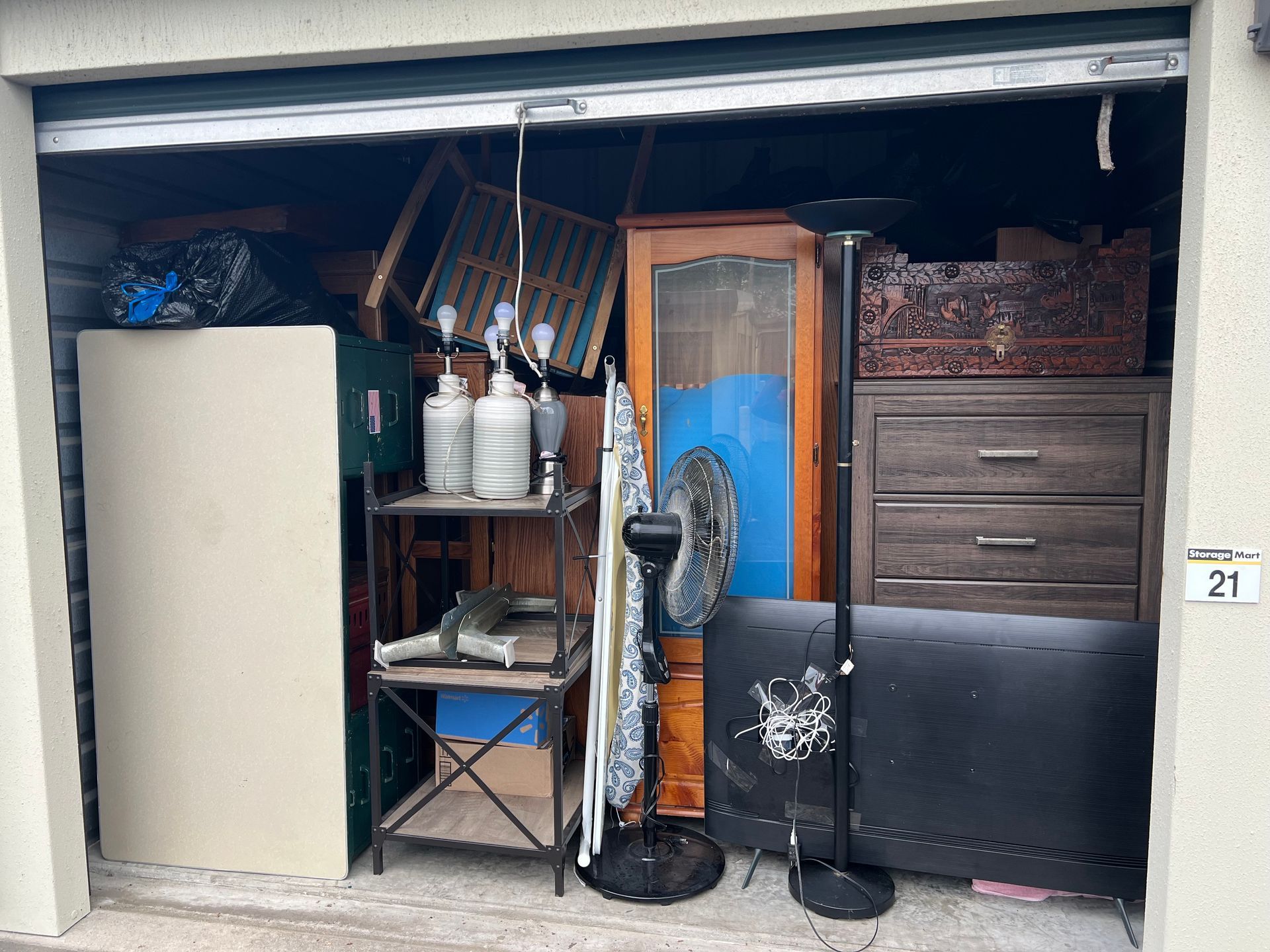 Storage unit packed with furniture, appliances, and boxes; various shades of brown, black, and metal.
