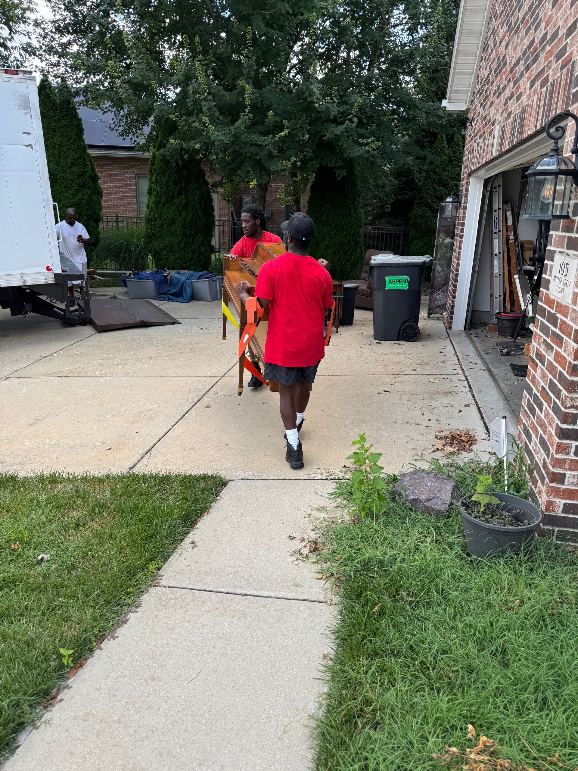 Two men moving furniture with a dolly near a brick garage; moving truck in background.