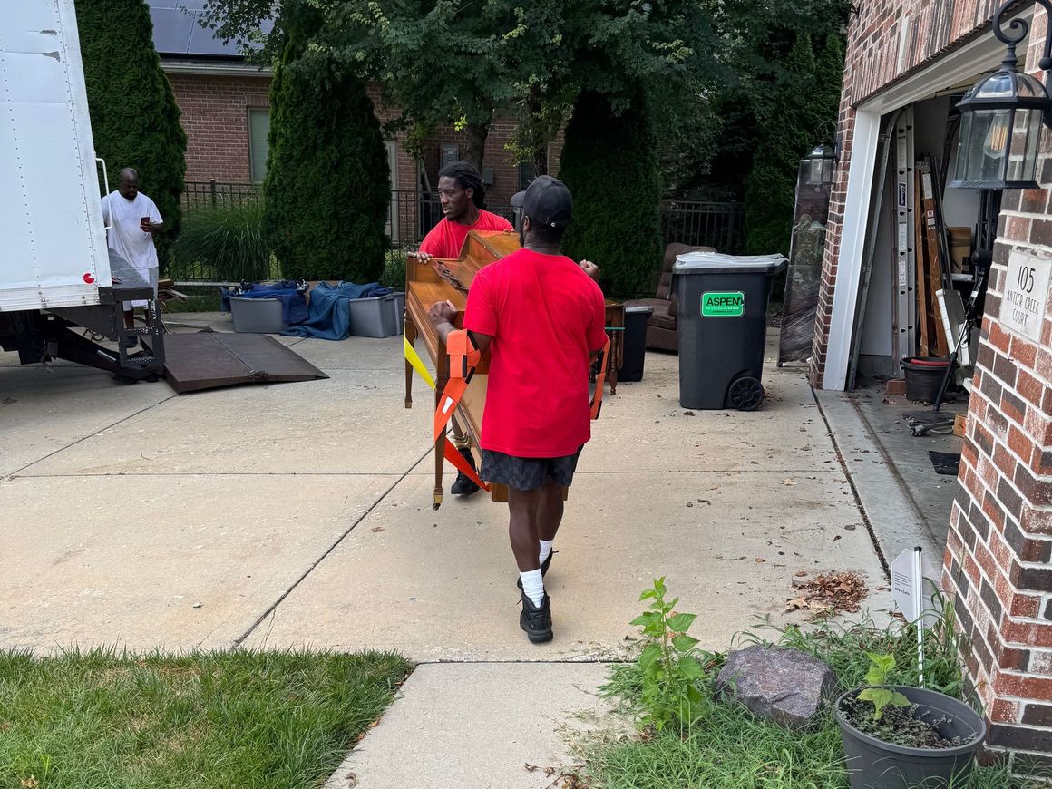 Three people moving furniture from a truck to a garage, outside a house; two are carrying a wooden chair.