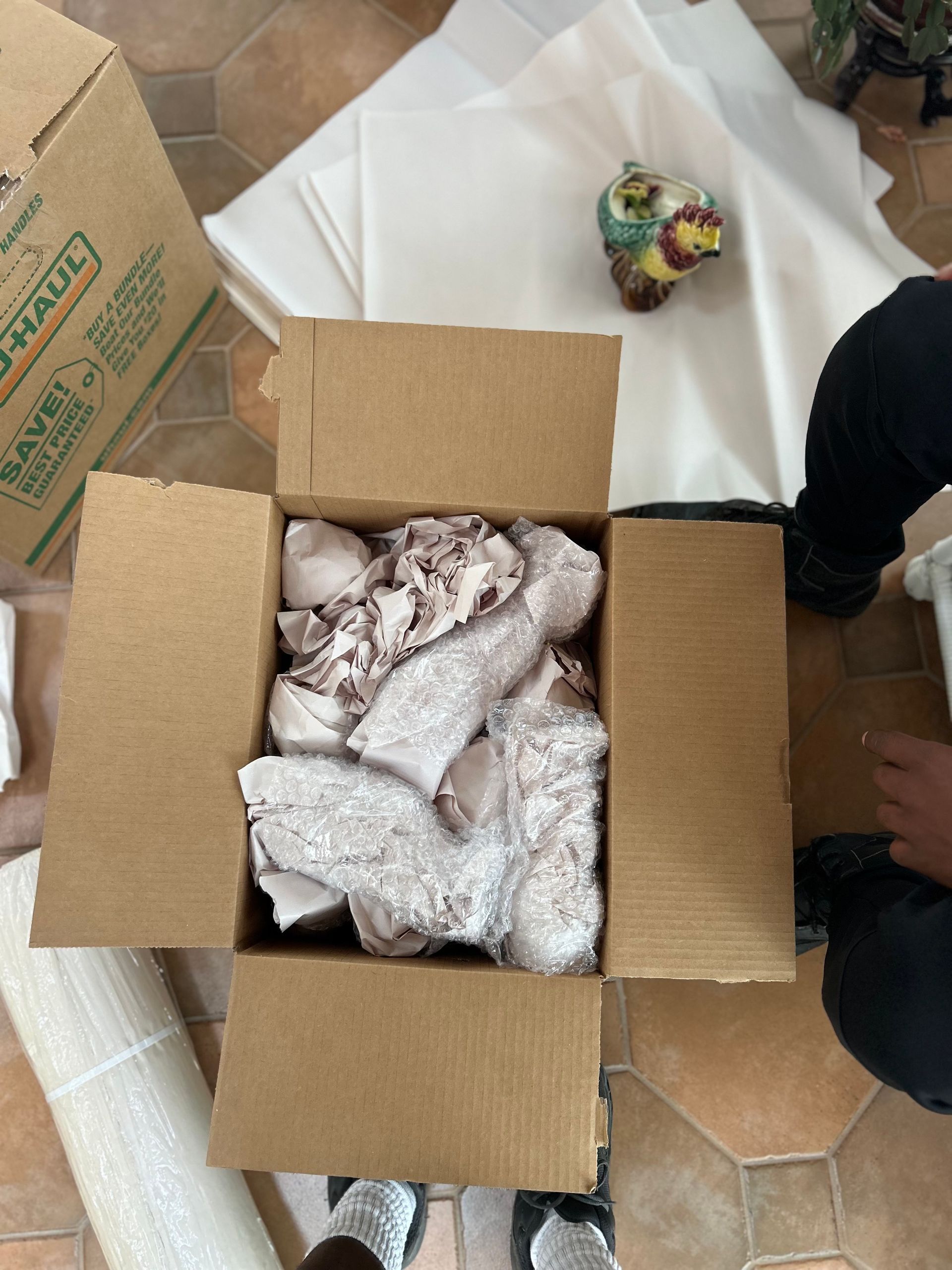 Open cardboard box with packing paper, surrounded by packing supplies and a figurine on a tiled floor.