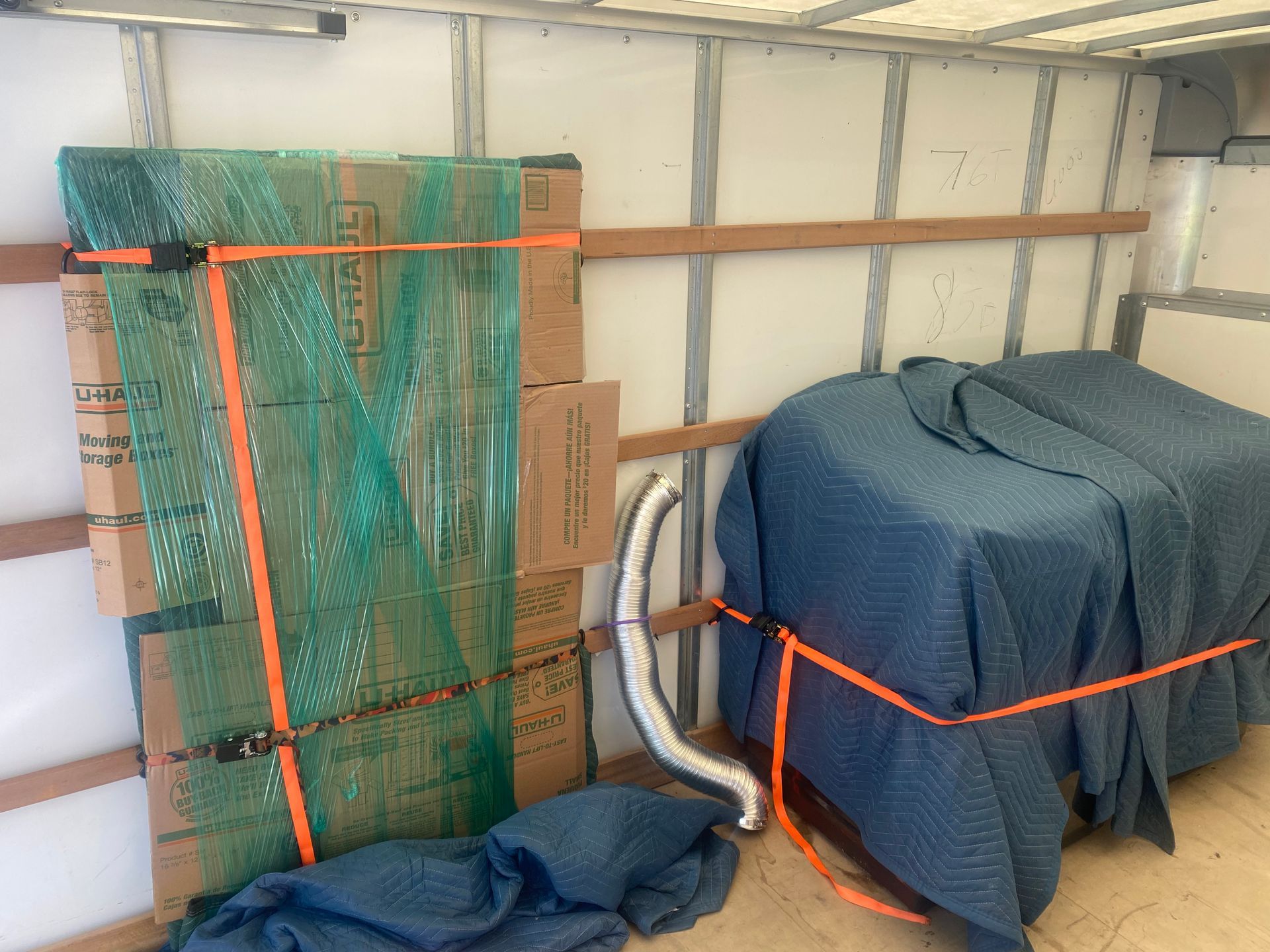 Boxes wrapped in green plastic and furniture covered in blue pads secured with orange straps inside a moving truck.