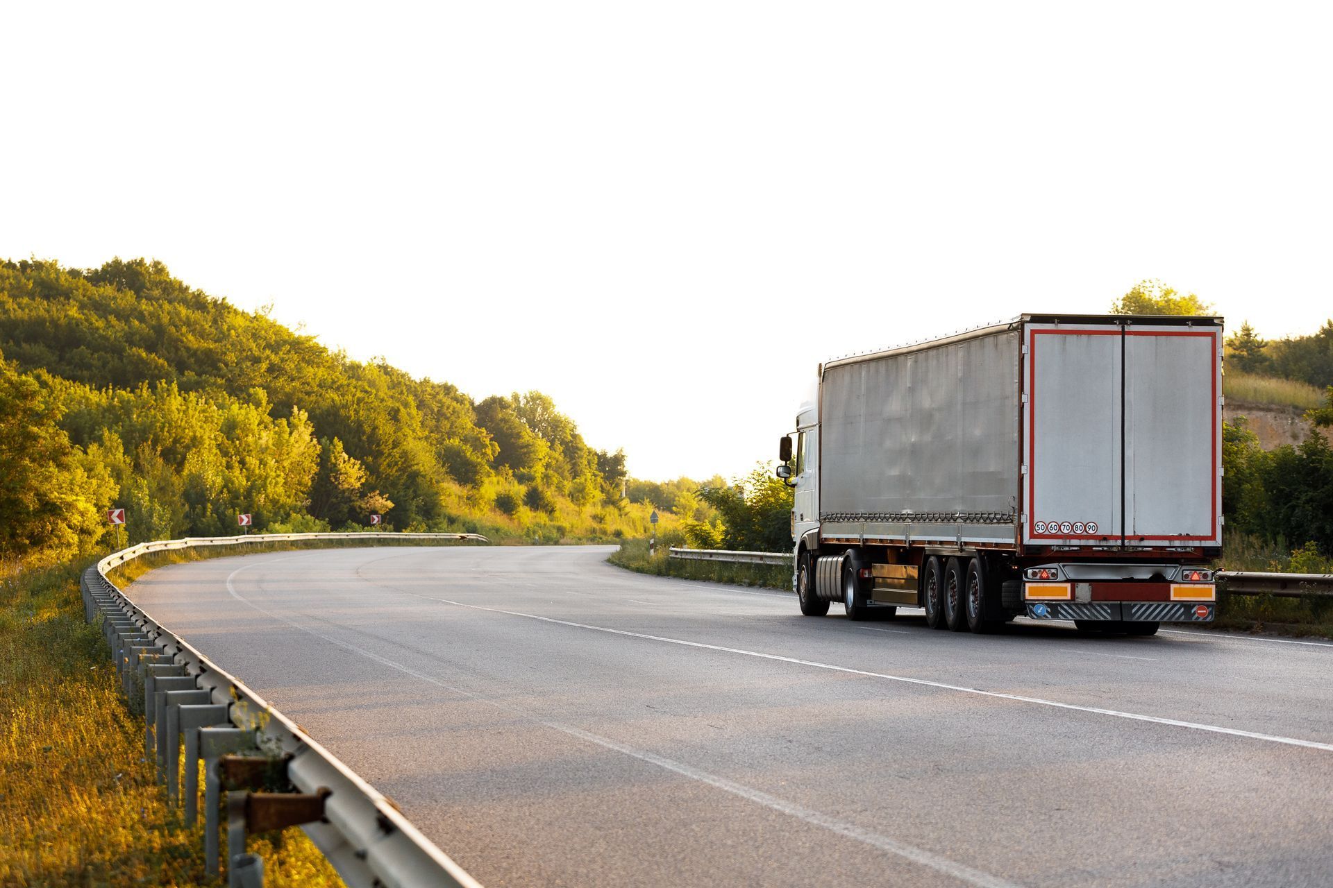 Semi-truck driving on a winding road through a sunlit, wooded landscape.