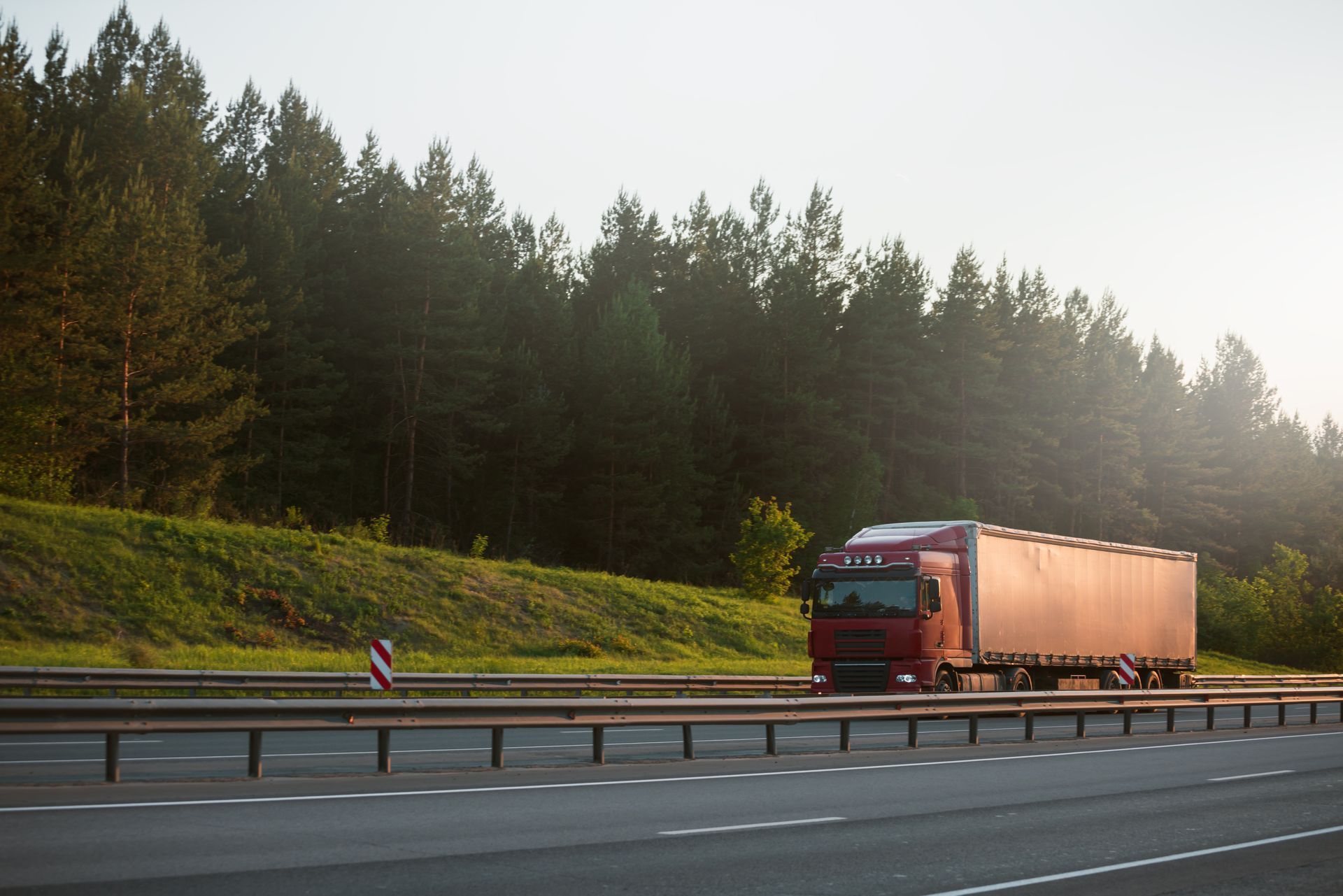 Red semi-truck driving on highway, alongside a treeline, under a bright sky.