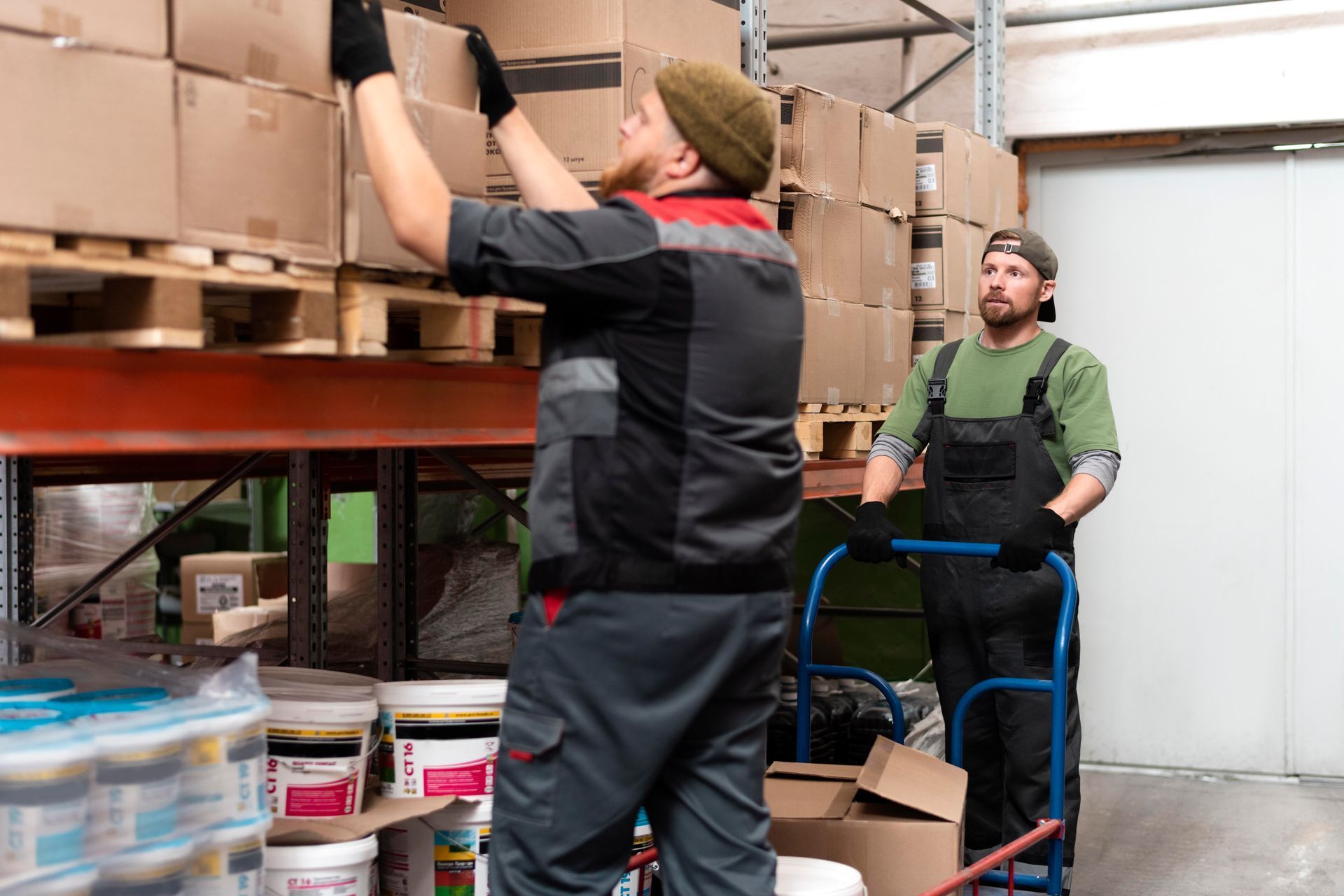 Two workers in a warehouse: one reaching for a box on a shelf, the other with a hand truck.