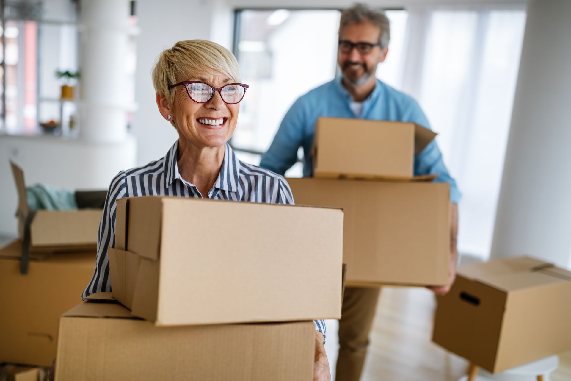 Smiling older couple carrying moving boxes in a bright room.