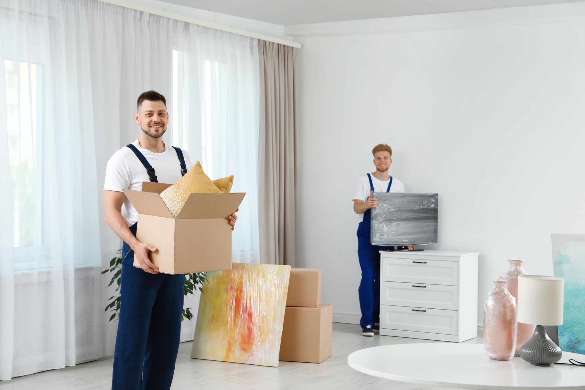 Two movers in a room: one holding a box, the other near a dresser. Boxes, art, and a table are visible.