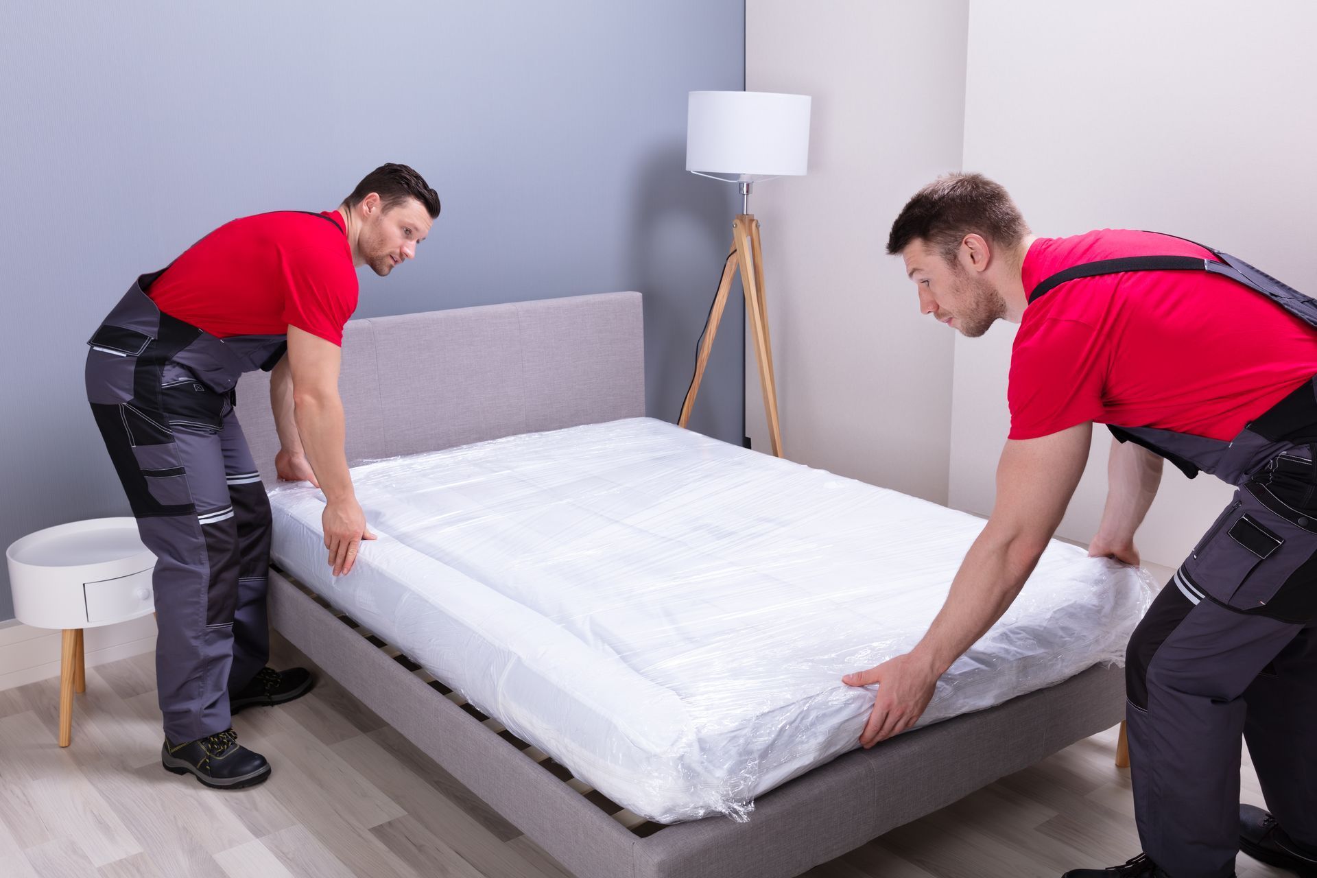 Two movers in red shirts and gray overalls lifting a wrapped mattress onto a bed frame.