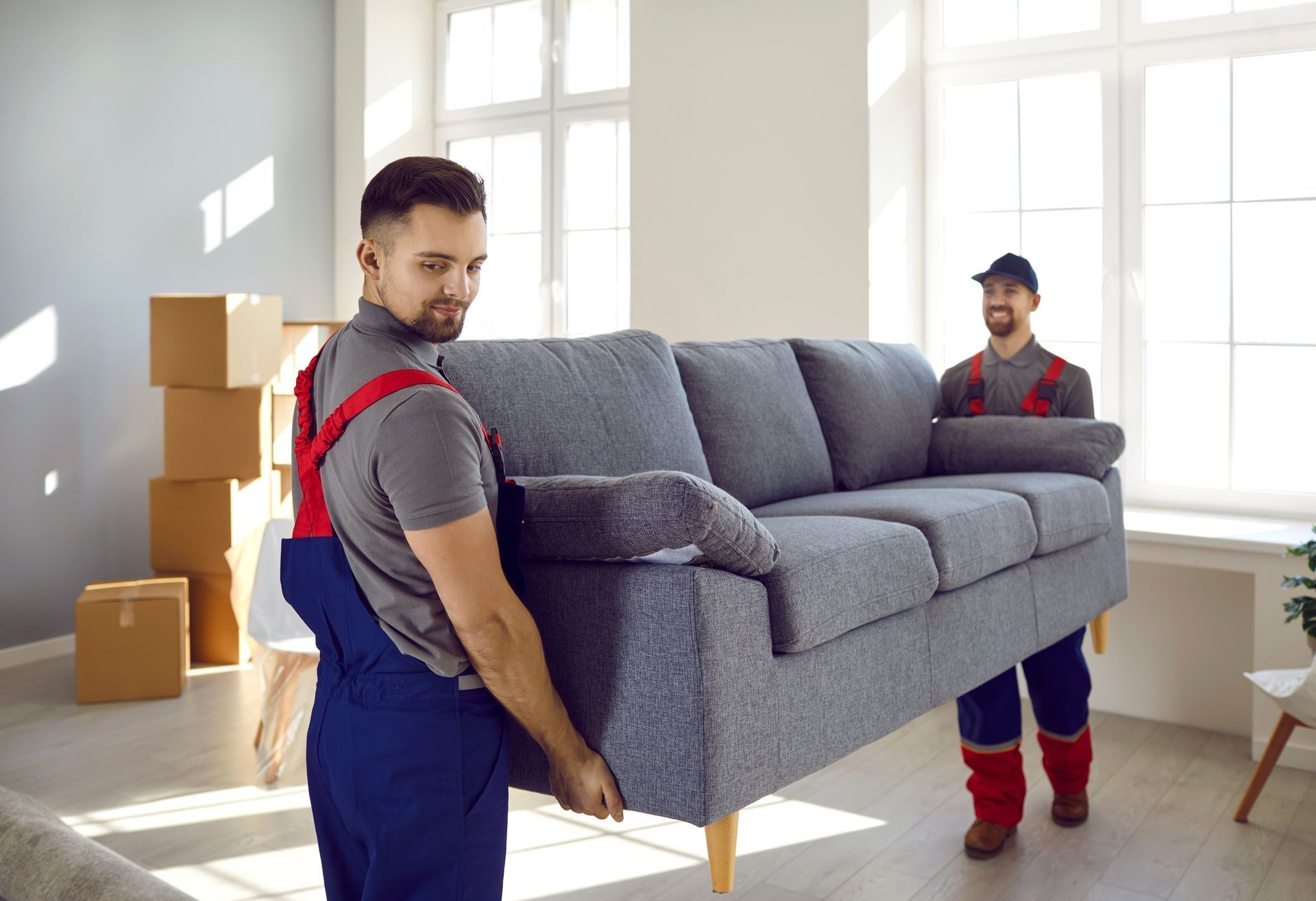 Two movers in overalls carry a gray couch in a well-lit room with boxes and a window.