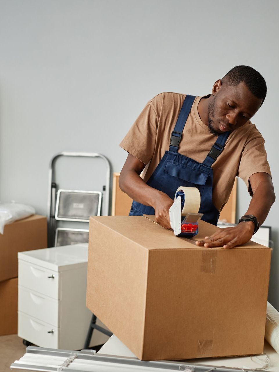 Man in overalls seals cardboard box with packing tape. Boxes, white cabinet, and ladder in room.