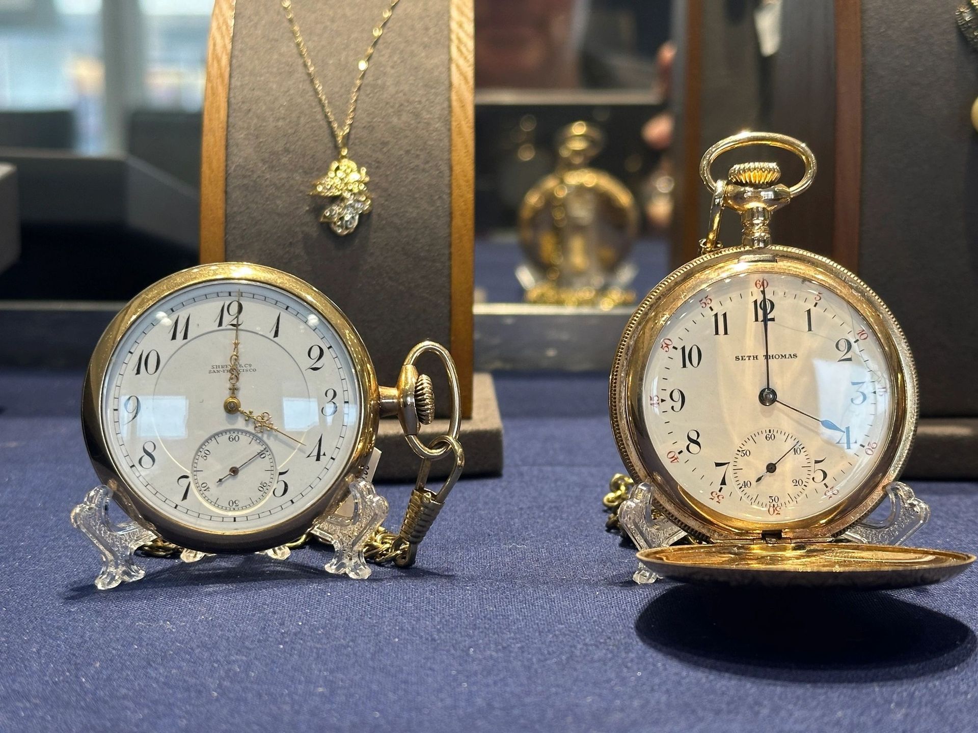 Two ornate gold pocket watches on stands displayed on a blue surface, with a gold necklace in the blurred background.