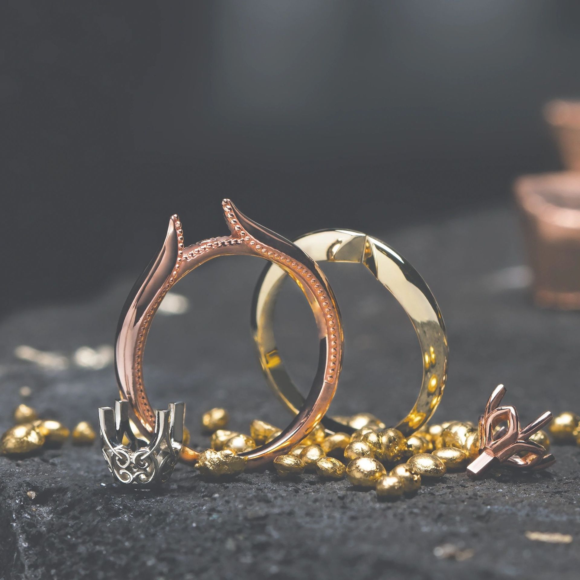 Two rose gold and yellow gold ring bands rest on a dark, textured surface alongside loose gold metal bits.