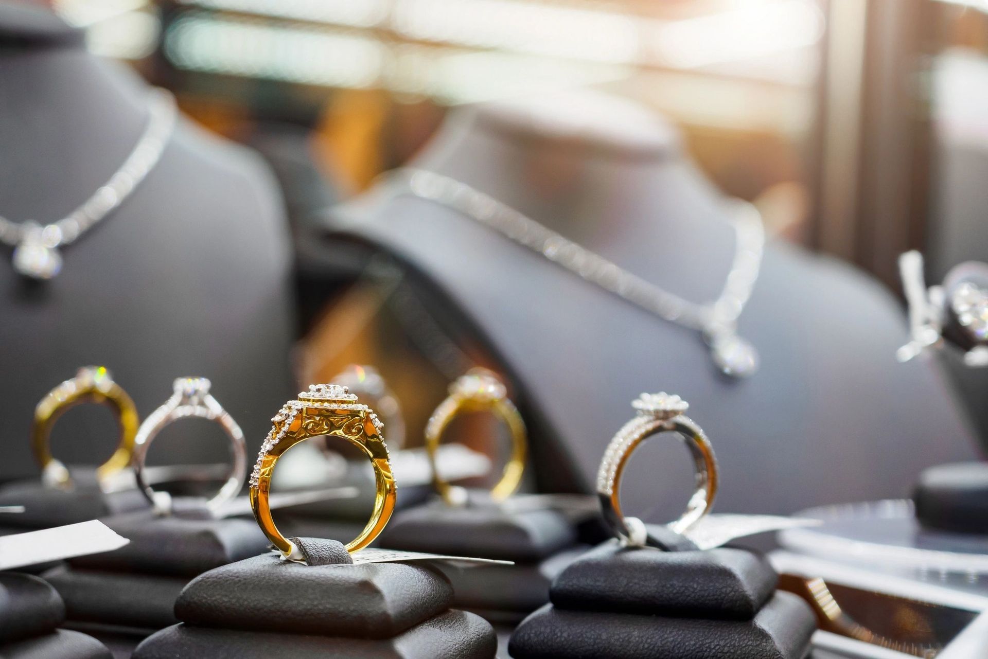 Gold and diamond rings displayed on velvet stands with necklaces on busts in a blurred background.