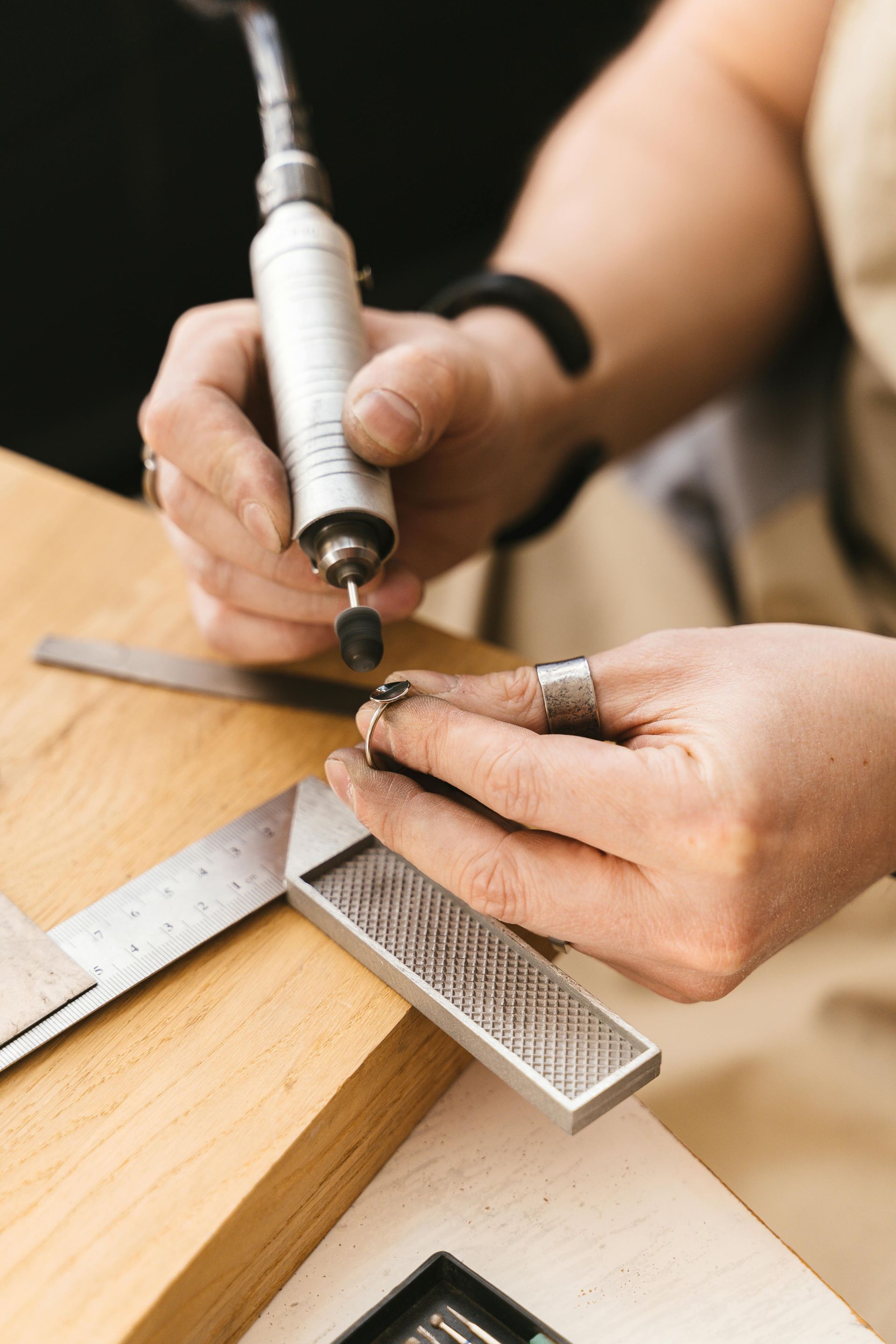 A jeweler uses a handheld rotary tool to polish or refine a small ring on a wooden workbench with a metal square nearby.